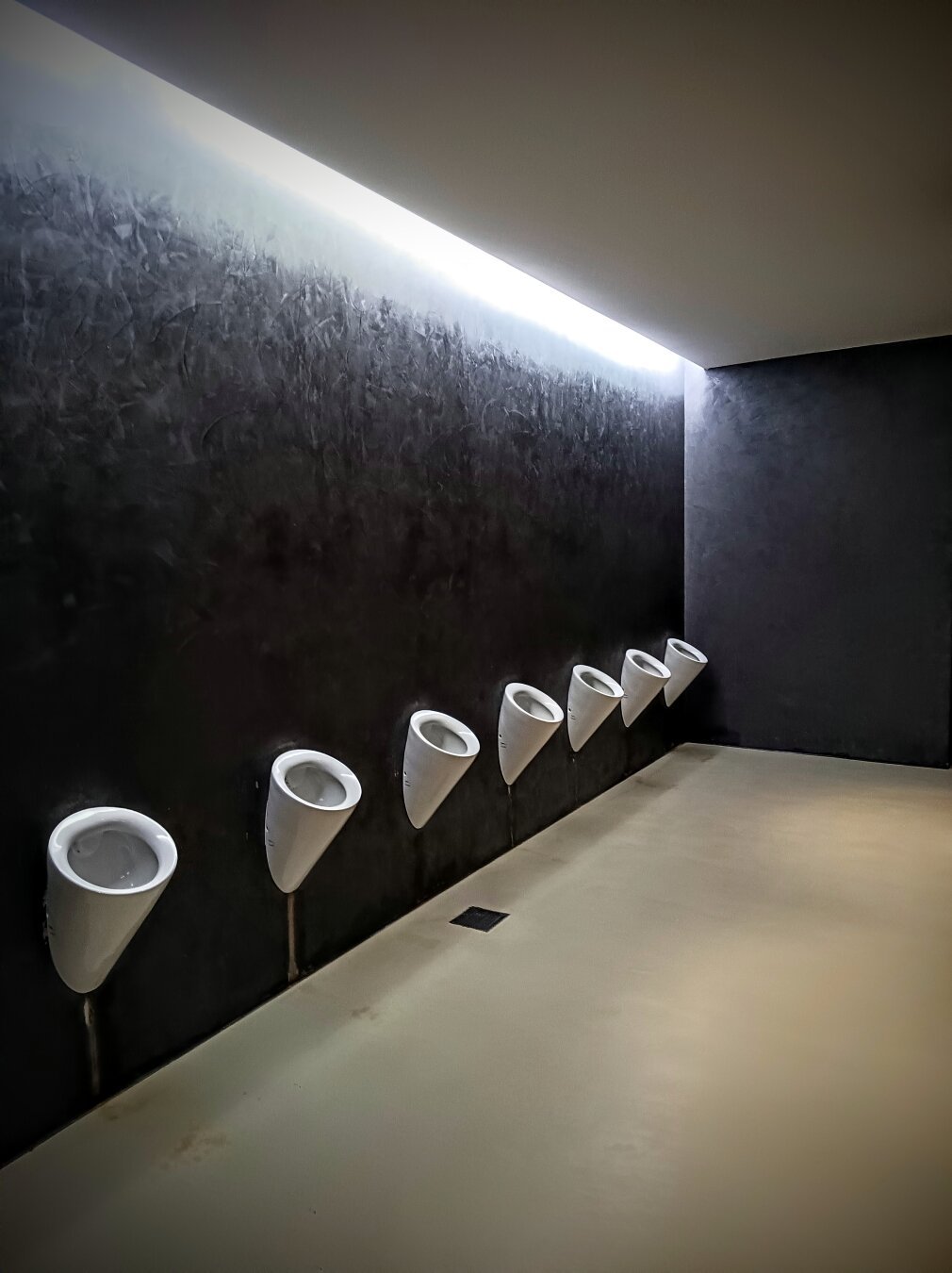 A modern men's restroom with eight white urinals mounted in a row along a dark, textured wall. A narrow strip of overhead lighting creates a dramatic contrast, and the floor is a smooth, light-colored surface. The overall design is sleek and minimalist.