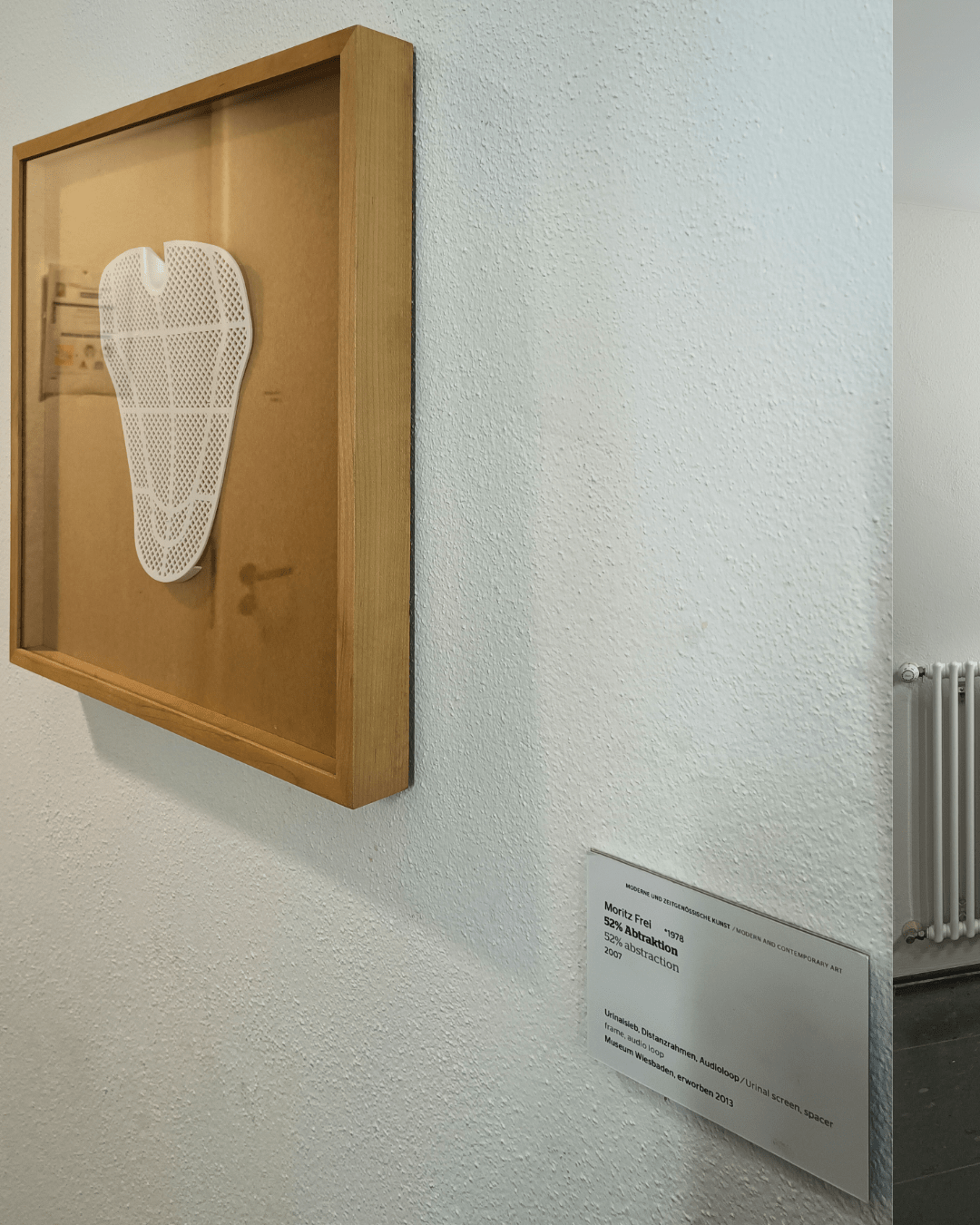 A framed artwork featuring a white perforated urinal screen mounted on a beige background. Below the frame is a plaque with the title "52% Abstraction" by Moritz Frei, mentioning its acquisition by the Museum Wiesbaden in 2013.