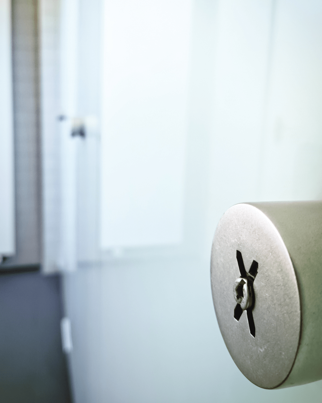 Close-up of a metal door handle with an “X” design on a blueish glass bathroom stall, with the background out of focus showing tiled walls and a white radiator.
