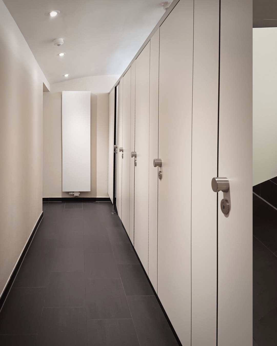 A clean, modern restroom with five closed stall doors on the right and a tall white radiator at the end of a dark-tiled hallway.