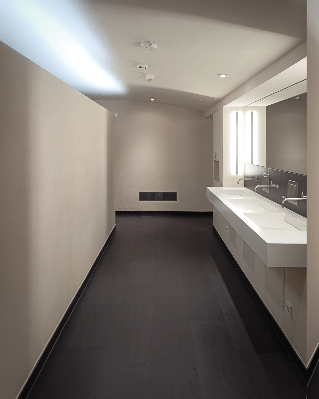 A modern, minimalistic public restroom with two white sinks, mirrors, and dark floor tiles against light-colored walls.