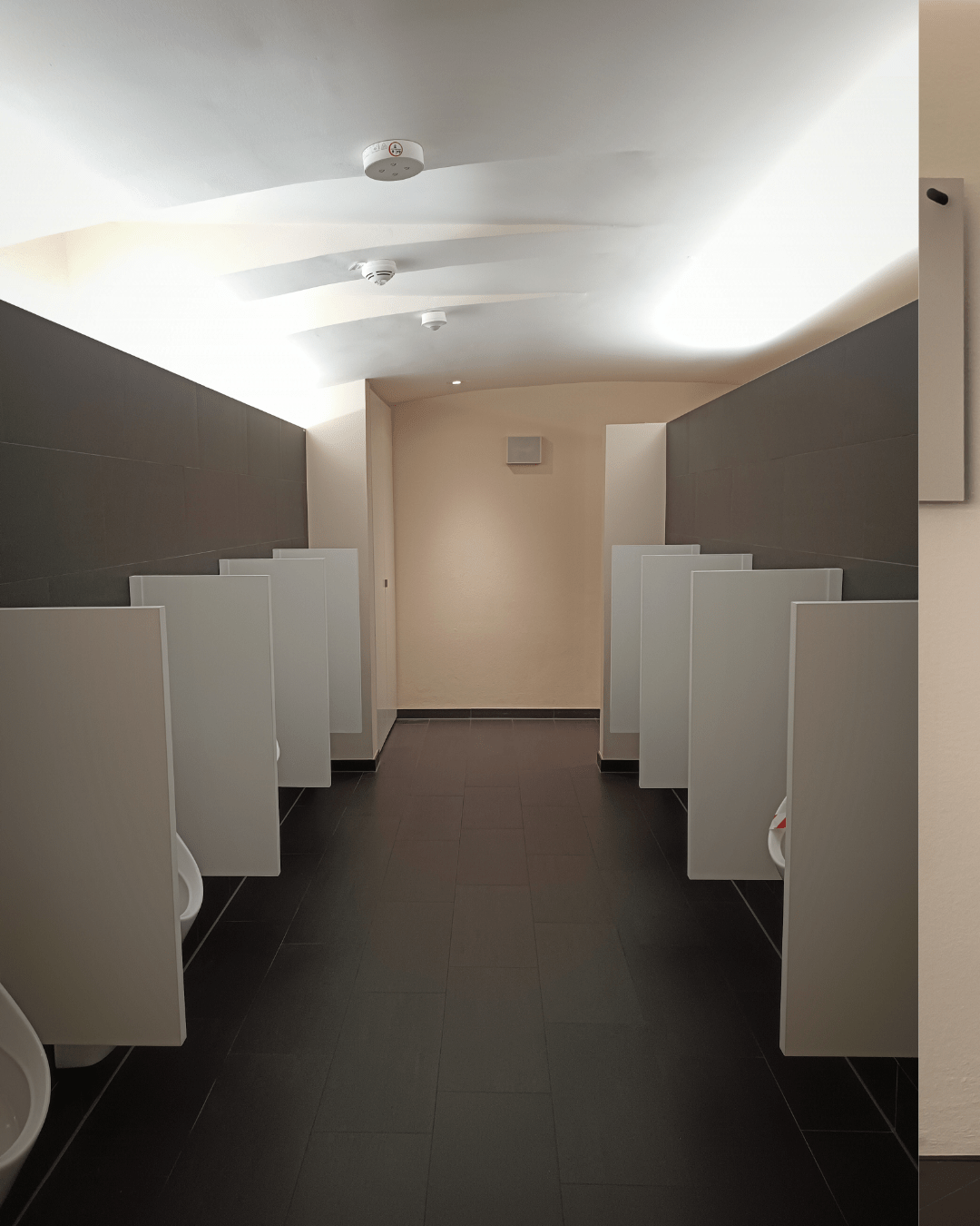 Interior of a modern men's restroom featuring white urinals against dark walls and tiles underneath and a well-lit ceiling.