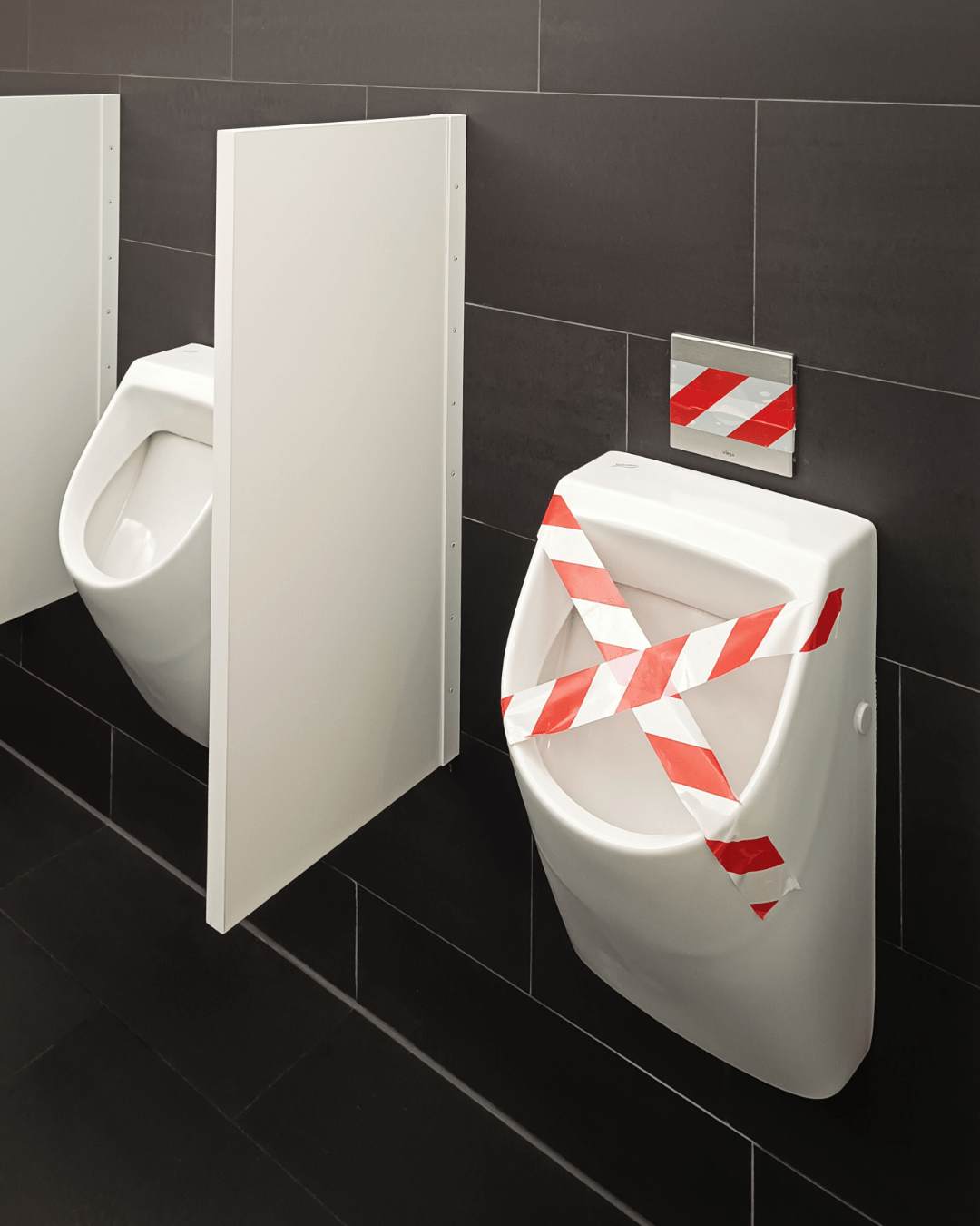Two urinals in a restroom with one blocked off by white and red striped tape.