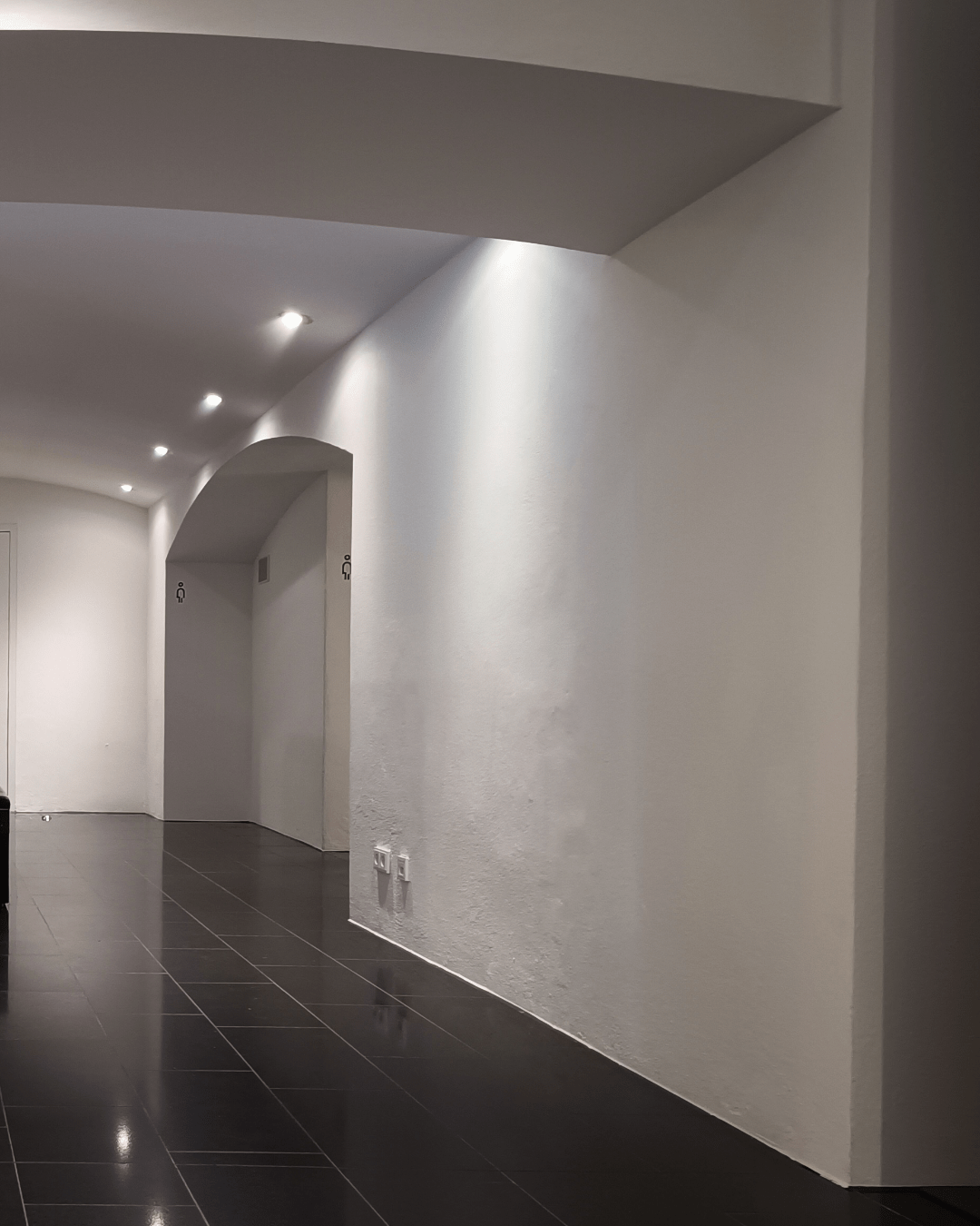 A spacious, modern hallway (barrel vault) with white walls and black tiled flooring, featuring multiple doorways, recessed lighting and a dark wooden divider construction wit integrated seating in the center. Pictograms indicate men's bathroom on the right and women's bathroom, baby change, disabled bathroom on the left.

(image part 2/2)