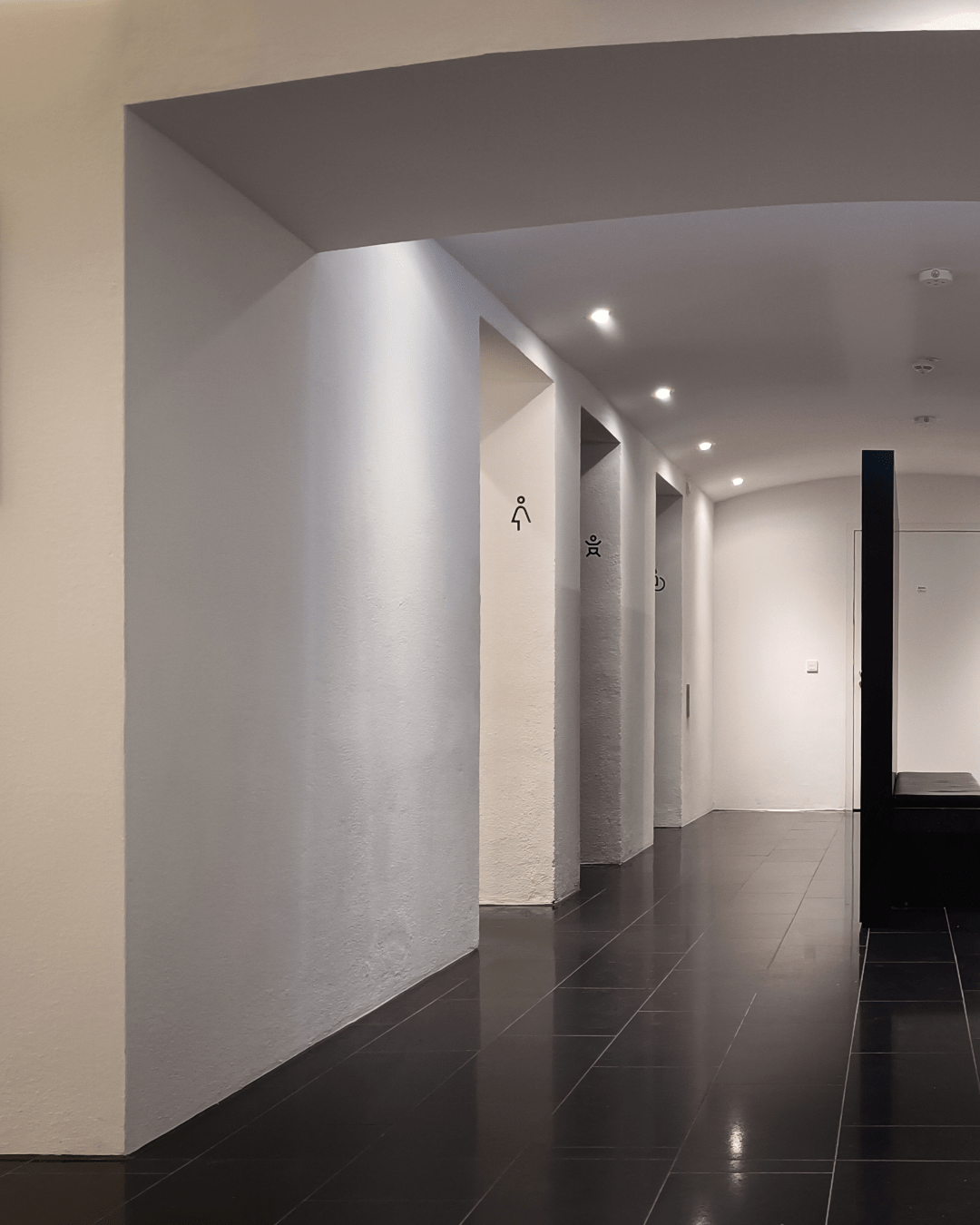 A spacious, modern hallway (barrel vault) with white walls and black tiled flooring, featuring multiple doorways, recessed lighting and a dark wooden divider construction wit integrated seating in the center. Pictograms indicate men's bathroom on the right and women's bathroom, baby change, disabled bathroom on the left.

(image part 1/2)