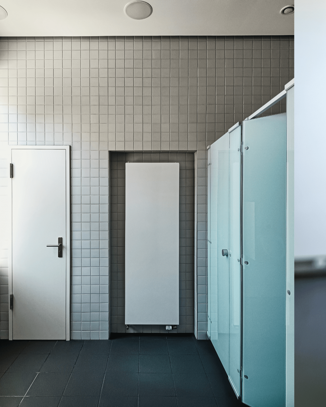 A clean public restroom with blueish frosted glass cubicles on the right, a white radiator in the center, and a plain white door on the left. The walls are tiled in light grey.