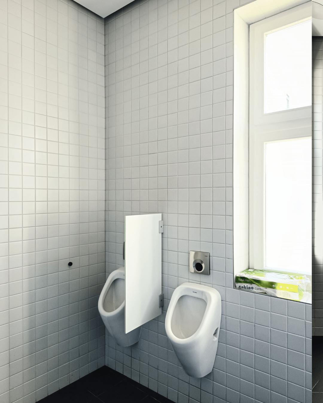 Two white urinals in the corner of a tiled bathroom, separated by a white privacy panel. A pack of toilet paper sits on the window ledge beside them.