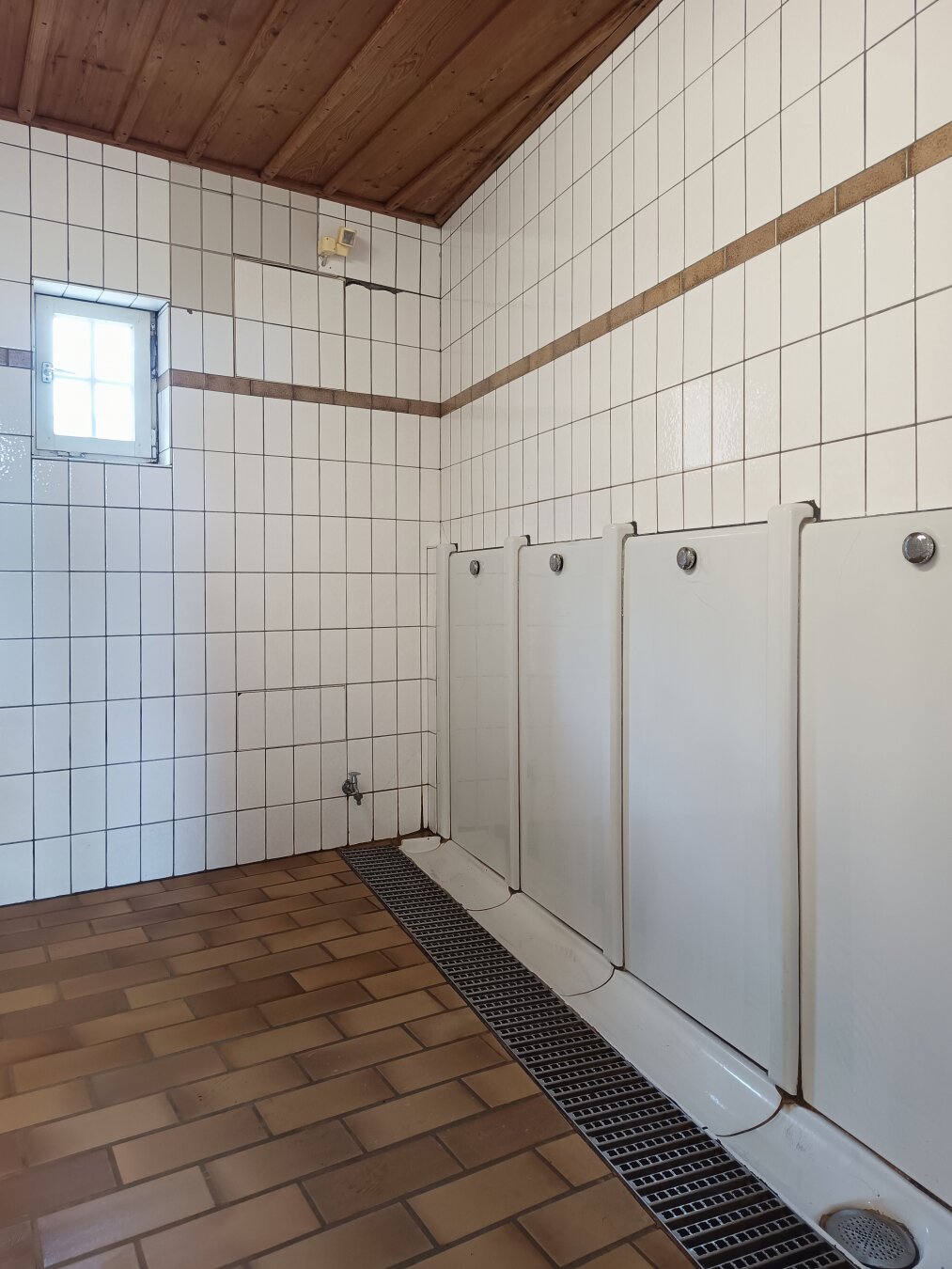 White-tiled restroom with three urinals against the wall, a window allowing light in, and a brown tiled floor with a drain. The atmosphere is clean and utilitarian.