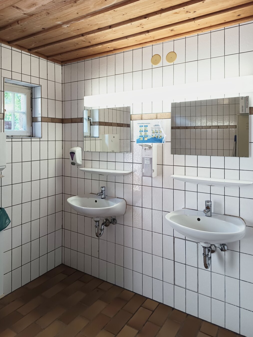 Two white sinks with mirrors in a tiled bathroom with wooden ceiling. A soap dispenser is on the left, and a poster is above the sinks. Bright, clean atmosphere.