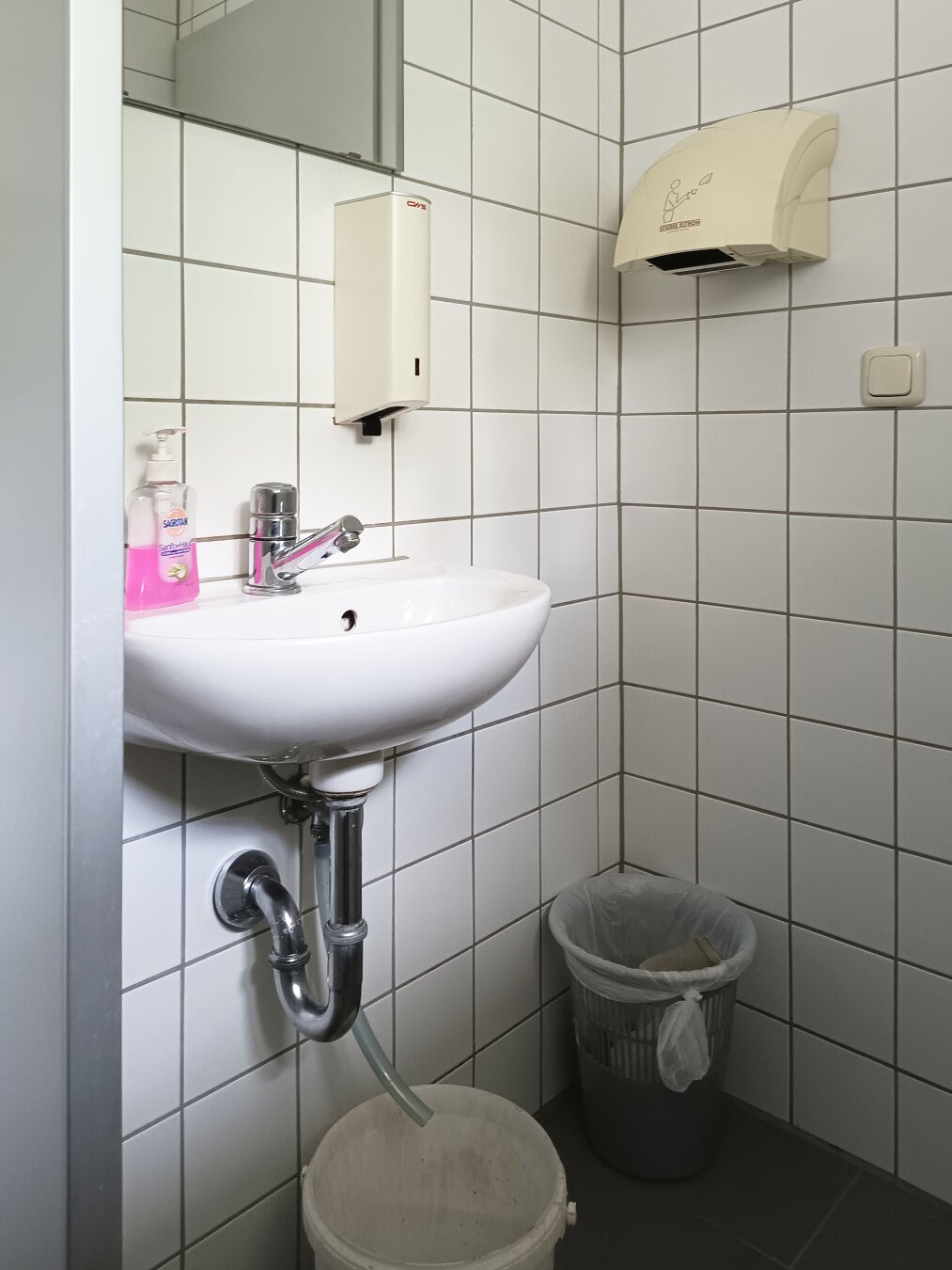 Small bathroom with white tiled walls, featuring a white sink, chrome faucet, hand soap, paper towel dispenser, and a trash bin. A bucket is placed under a short tube connected to the water outlet underneath the sink. Clean and utilitarian ambiance.