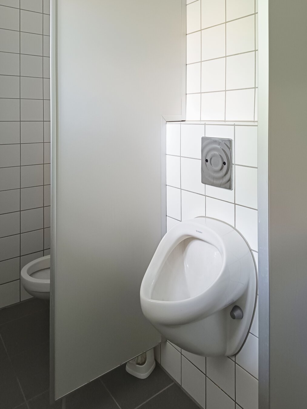 A clean, tiled restroom features a white urinal mounted on the wall next to a toilet partially visible behind a partition. The mood is sterile and functional.