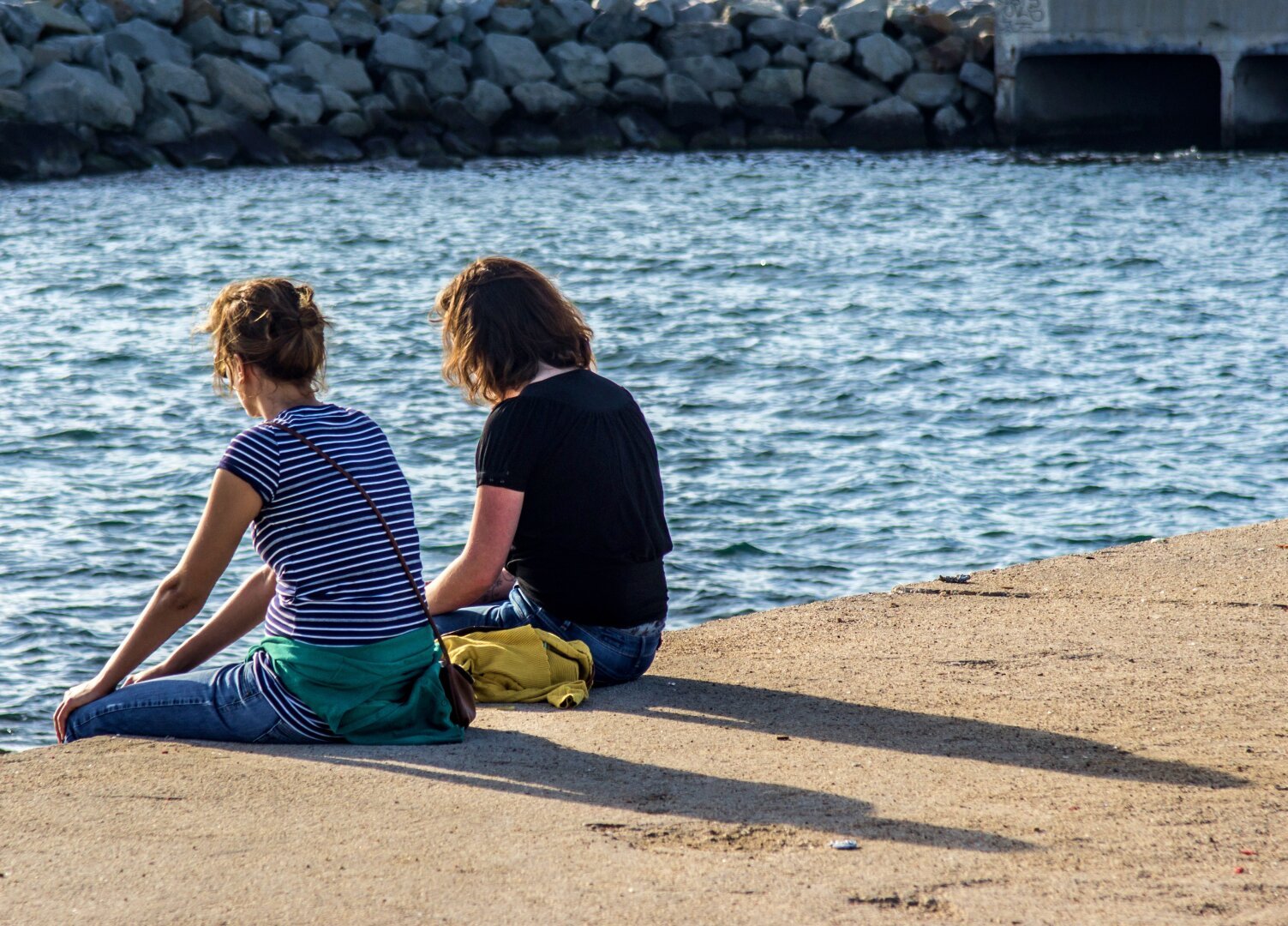 Se ven dos chicas de espaldas, mirando al mar en un puerto urbano proyectando unas alargadas sombras debido al sol fuerte de esa hora del mediodía.

Two girls are seen with their backs turned, looking at the sea in an urban port, casting long shadows due to the strong sun at that midday hour.