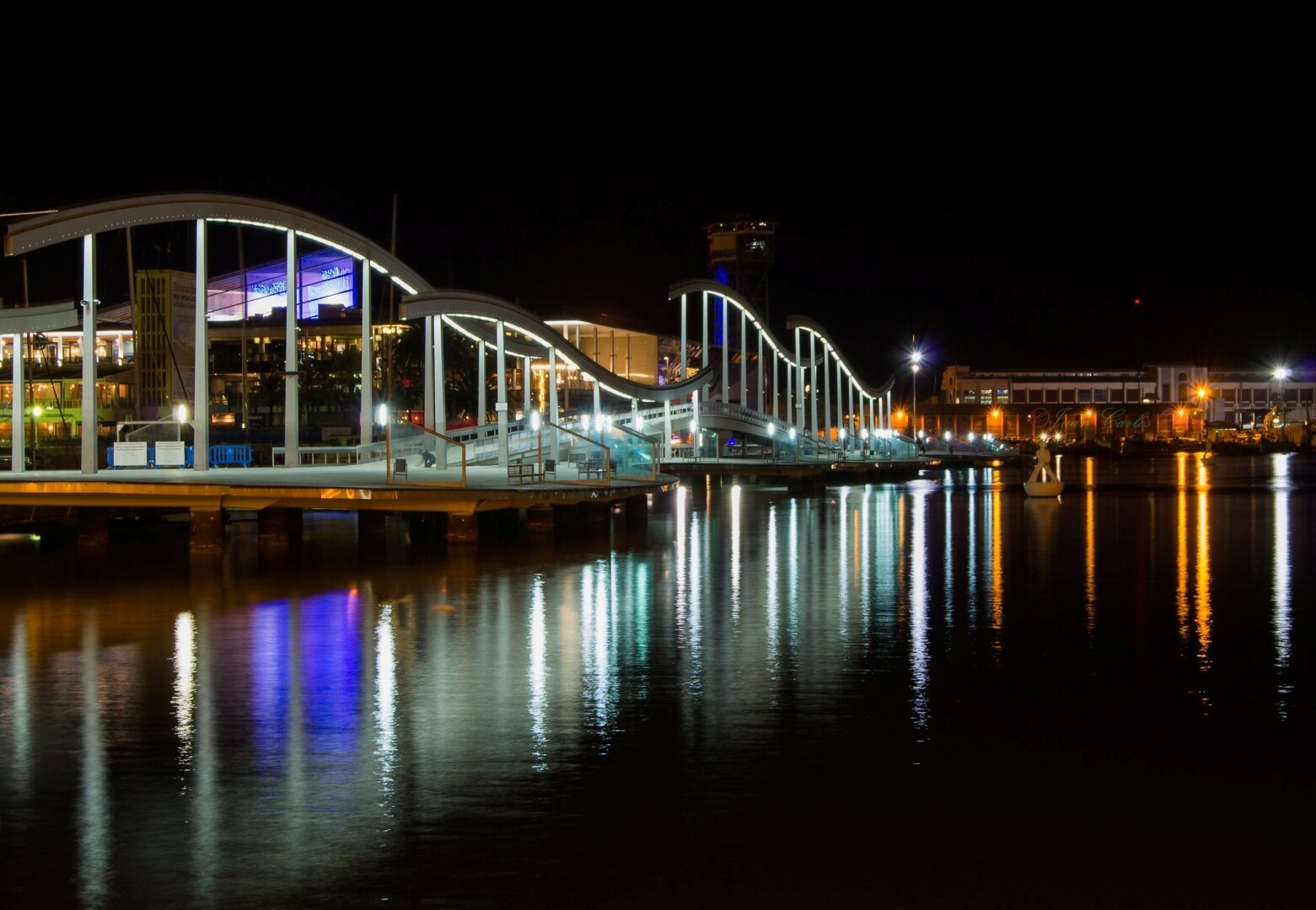 Una larga exposición nocturna del puente con los reflejos de las luces en el agua del mar del centro comercial Maremagnum, en Barcelona, España.

A long exposure night photo of the bridge with the reflections of the lights in the seawater of the Maremagnum shopping center, in Barcelona, Spain.