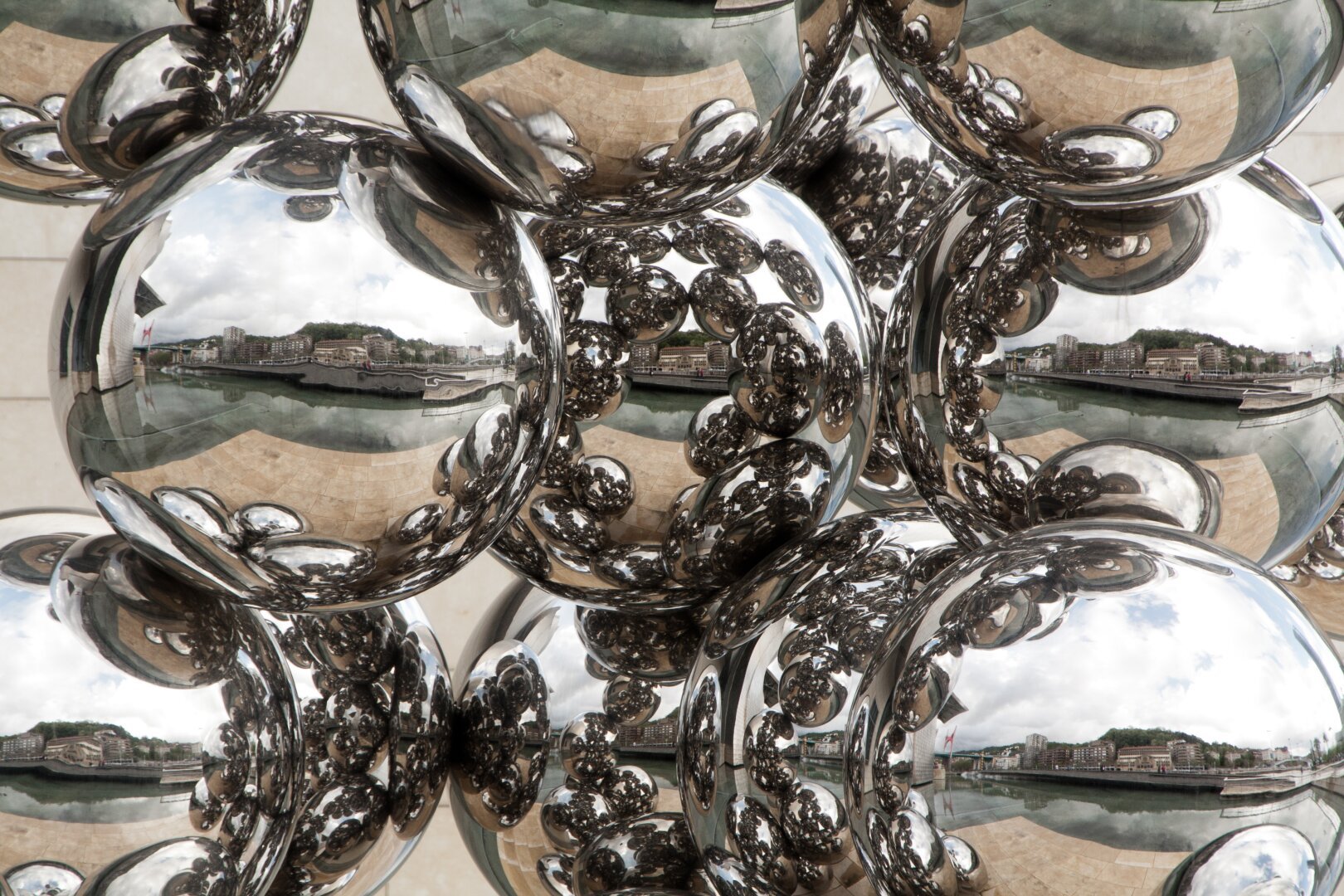 A close-up view of a mirrored stainless steel sculpture at the Guggenheim Museum in Bilbao, Spain. The reflective spheres capture distorted images of the surrounding cityscape, including modern buildings, a river, and the museum's exterior. The reflections create a mesmerizing kaleidoscopic effect, with multiple layers of repeating patterns. The cloudy sky and soft natural light enhance the metallic surface, making the reflections appear crisp and dynamic. The geometric arrangement of the spheres adds depth and dimension, drawing the viewer into an abstract yet immersive visual experience.