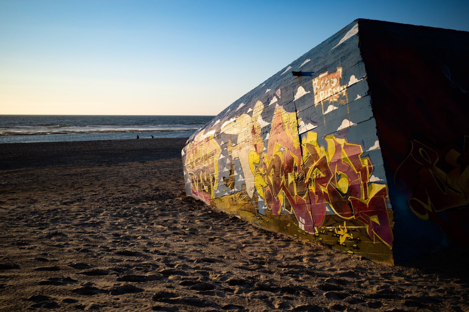 A graffiti-covered World War II bunker partially buried in the sand on a beach, illuminated by the warm golden light of the setting sun. The artwork on the concrete surface features vibrant yellow and red lettering, blending with a painted blue sky and white clouds. In the background, the calm sea stretches to the horizon, with two silhouetted figures sitting near the water’s edge. The scene captures a contrast between history, urban art, and nature.