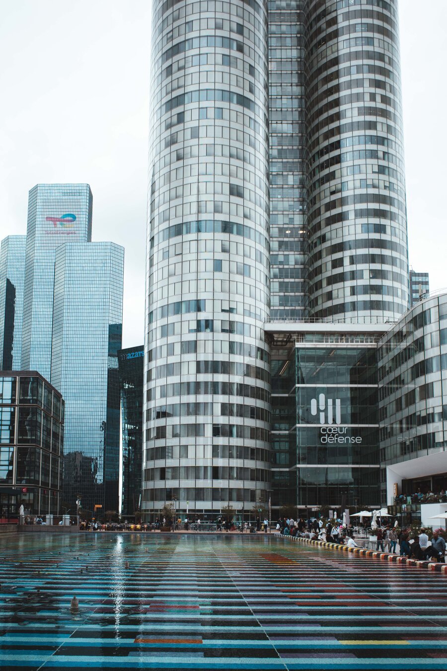 A modern architectural view of La Défense in Paris, featuring the glass and steel skyscrapers of Cœur Défense. The foreground showcases a colorful, reflective surface, with people sitting and walking around, adding a lively urban atmosphere.