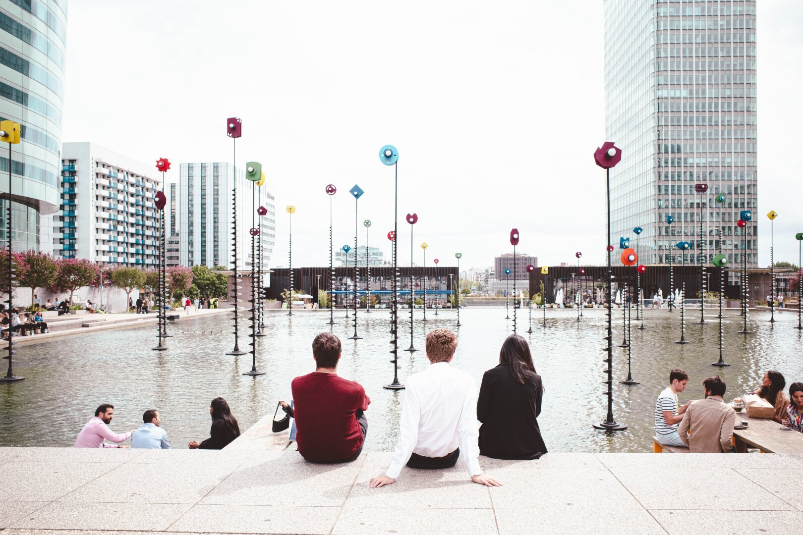 A group of three people sits on the edge of a large urban water feature, facing a modern cityscape with tall glass buildings. The water is adorned with numerous tall, black poles topped with colorful geometric shapes. Other people are seen socializing and relaxing around the area. The atmosphere is bright and lively, suggesting a popular public space in a metropolitan setting.