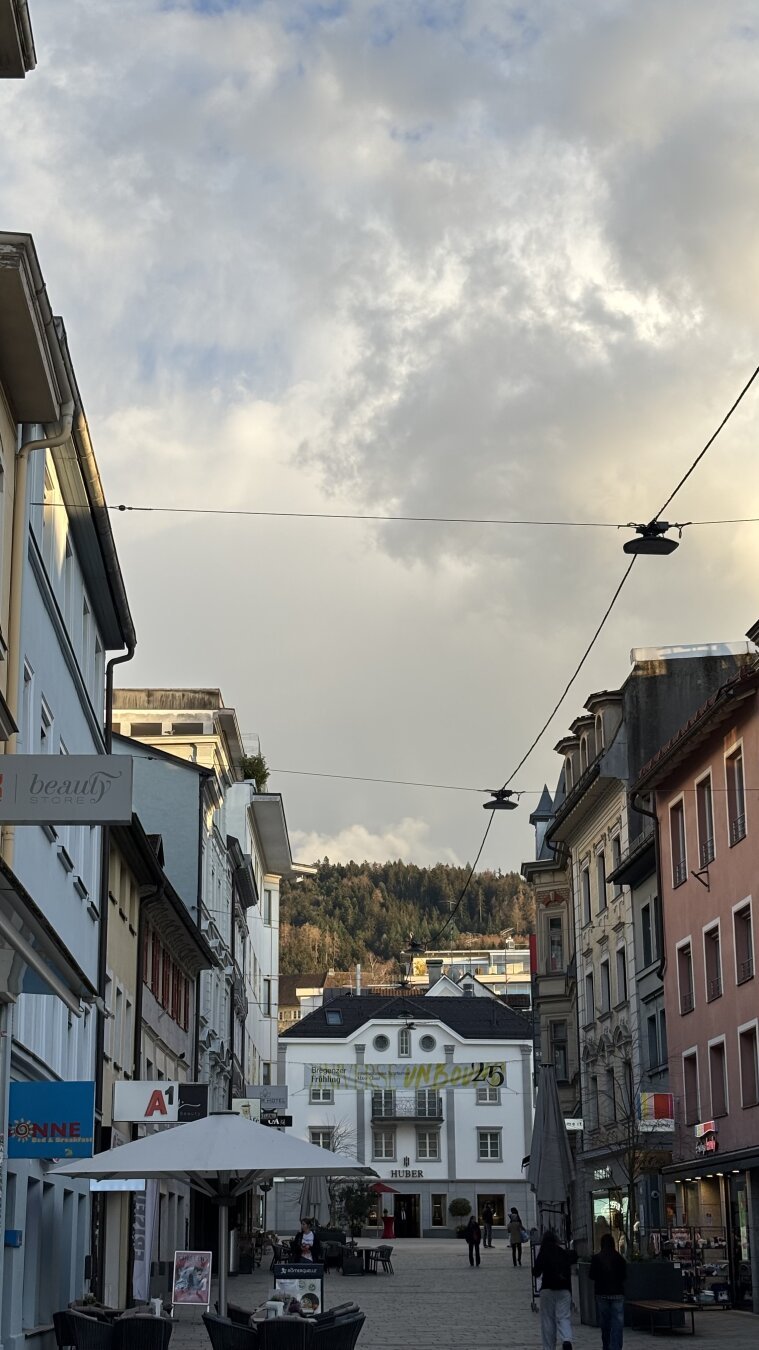 Die Fußgängerpassage in Bregenz. Links und rechts säumen Geschäfte die Kaiserstraße, geradezu läuft sie auf ein teureres Geschäft hinaus. Im Hintergrund fällt die Sonne auf den bewaldeten Pfänderhang, unberührt von den Wolken über der Straße.
