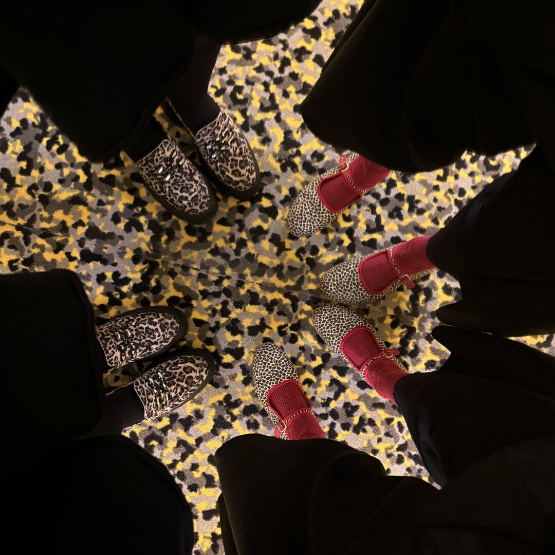 Zu sehen sind zwei Paar Füße in Schuhen im Leo-Look, gedoppelt durch einen Spiegel. Der Teppich hat ein wildes Punkte-Muster, des dem Muster der Schuhe ähnelt.