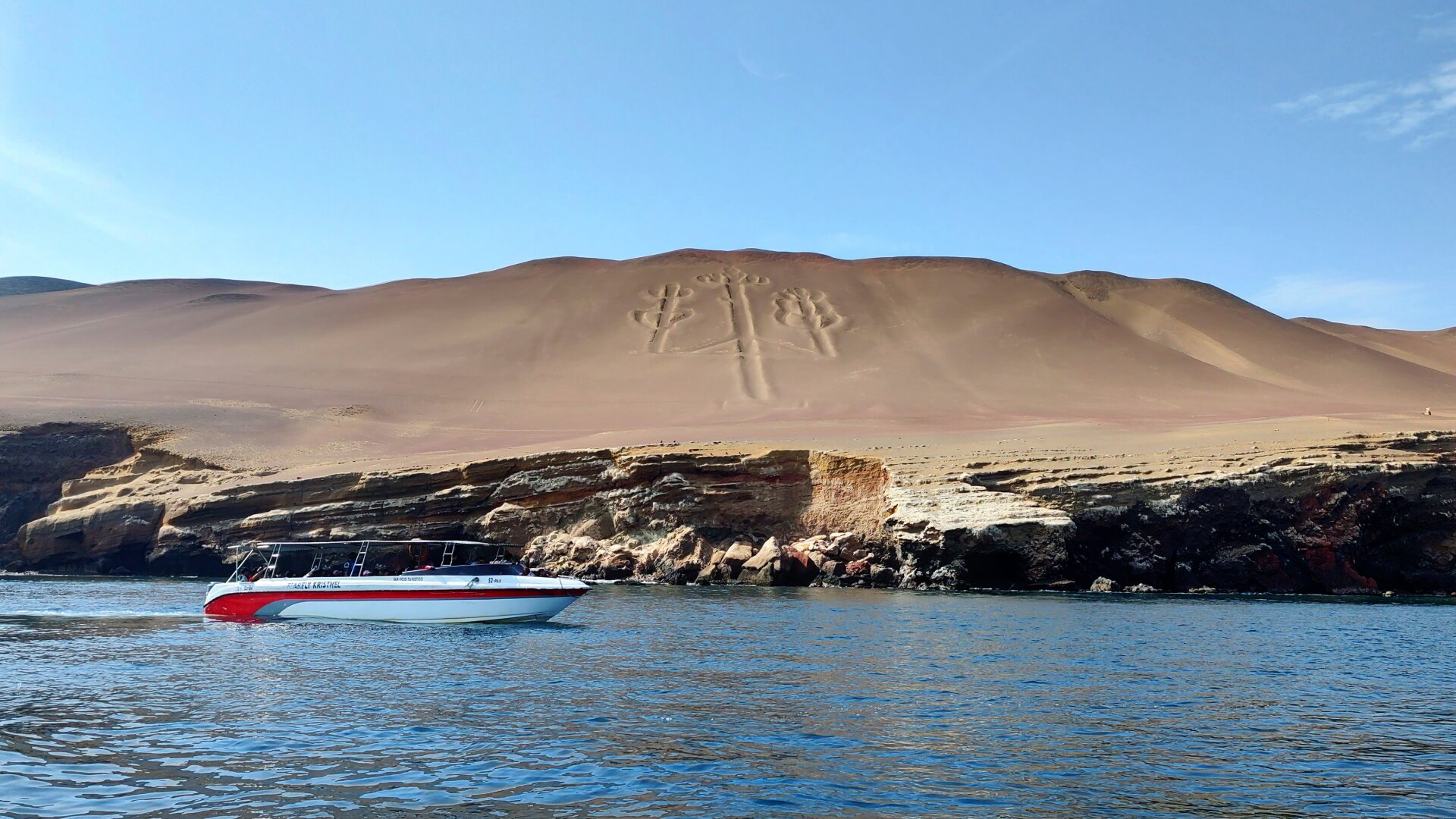 The candelabra of Paracas seen from the sea. Blue ocean in the foreground. A white and red boat enters the picture from the left. In the background the rocky cliffs. Their ochre colour merges directly into dune-like sand hills.
A large geometric figure is carved into the soft sand. It is reminiscentof a three-armed candlestick, or three ears of grain sharing a common stem.