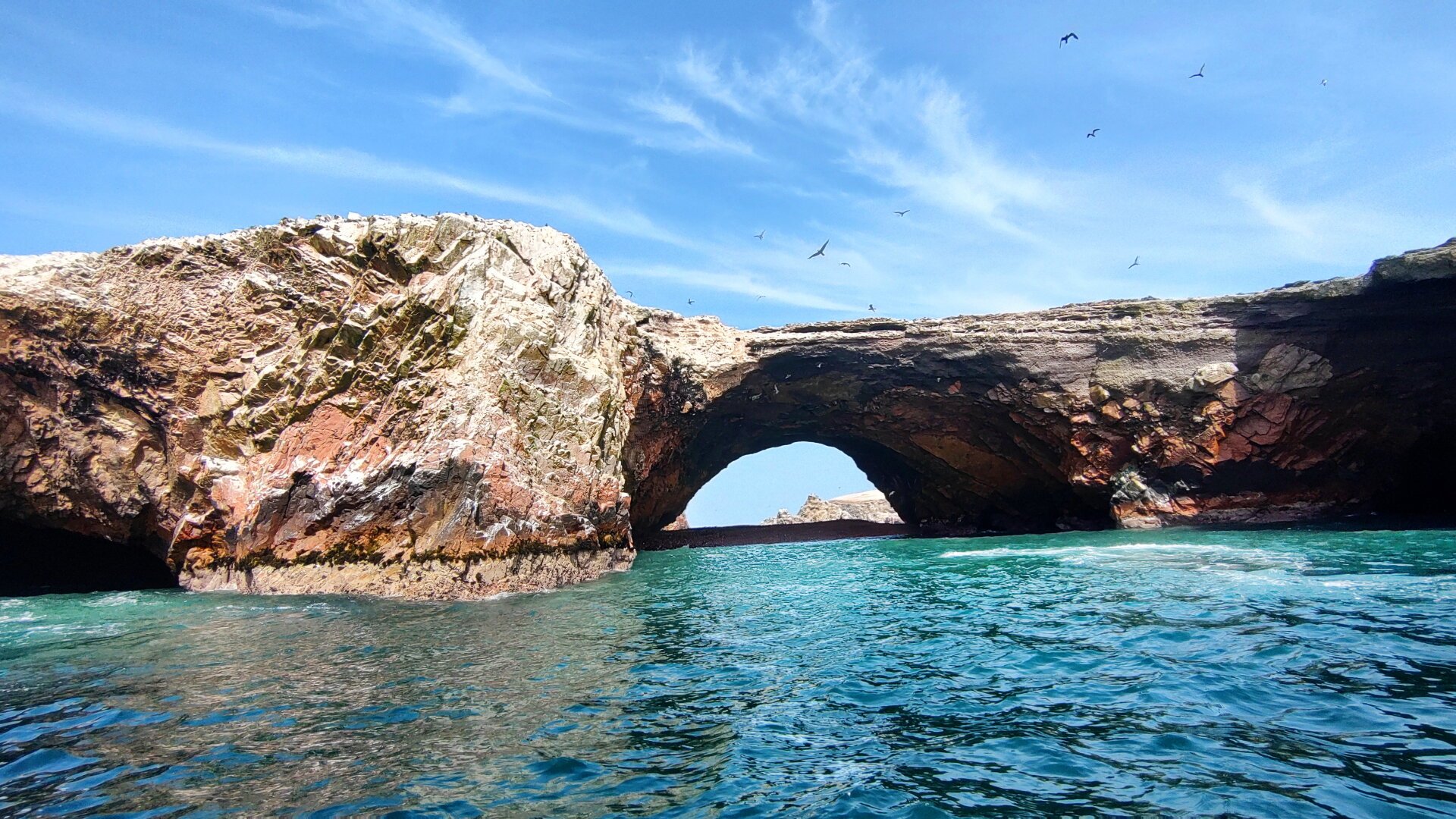 Turquoise water in the foreground. A rocky island. Cliffs shoot up steeply and form an almost levelled plateau. In the centre of the picture, an almost perfectly semi-circular arch cut out of the stone by wind and water, making the cliff look like a bridge.
Seabirds circle in the sky.