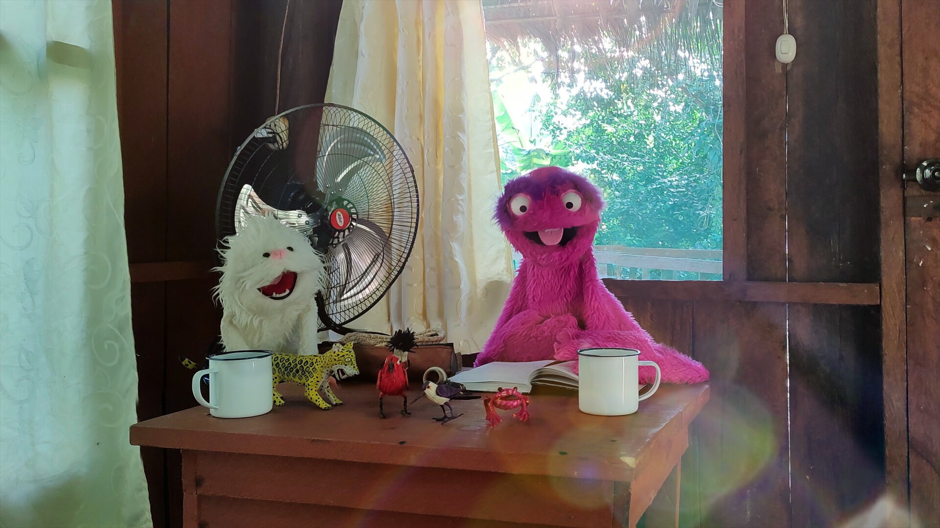 The inside of a simple wooden cabin. The sun shines in through the fly screen. You can see the green jungle outside. Sitting at the table are Adda, a small creature with fluffy white fur, and Garstl with his pink shaggy fur. In front of them is an open travel diary and handcrafted animal figurines: a jaguar, various birds and a toad.