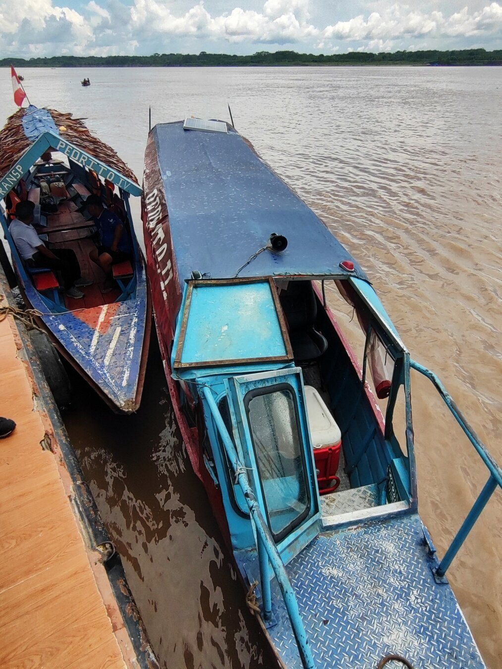 Two very narrow, elongated boats are moored on the river side of the ferry, one has a roof covered with palm leaves, the other has a blue tin
roof.