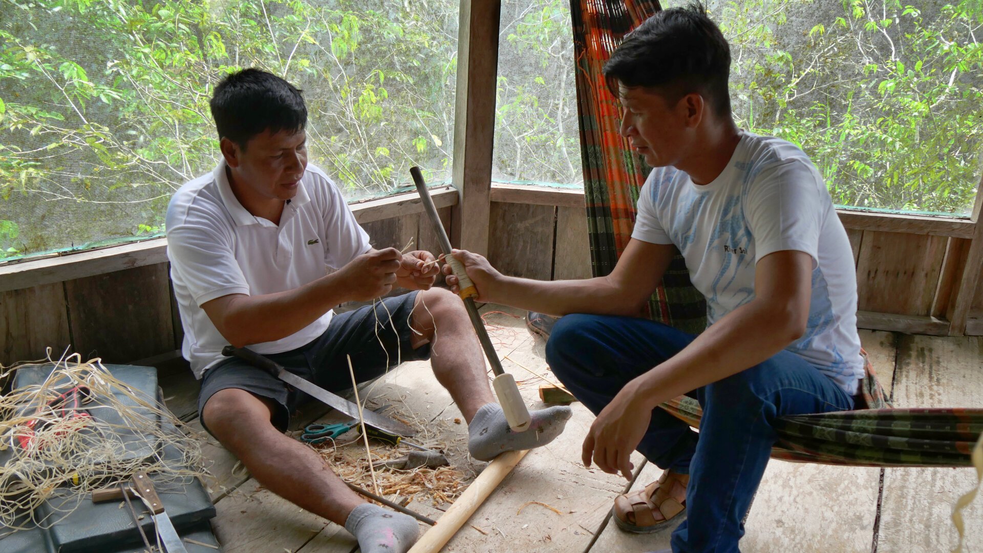 Two men are sitting in a corner of the hammock cabin. Surrounding them on the floor are tools, light-coloured wood shavings and natural-coloured raffia bast fibres. One of the men is holding a wooden blowpipe. The other one is wrapping raffia around the blowpipe.