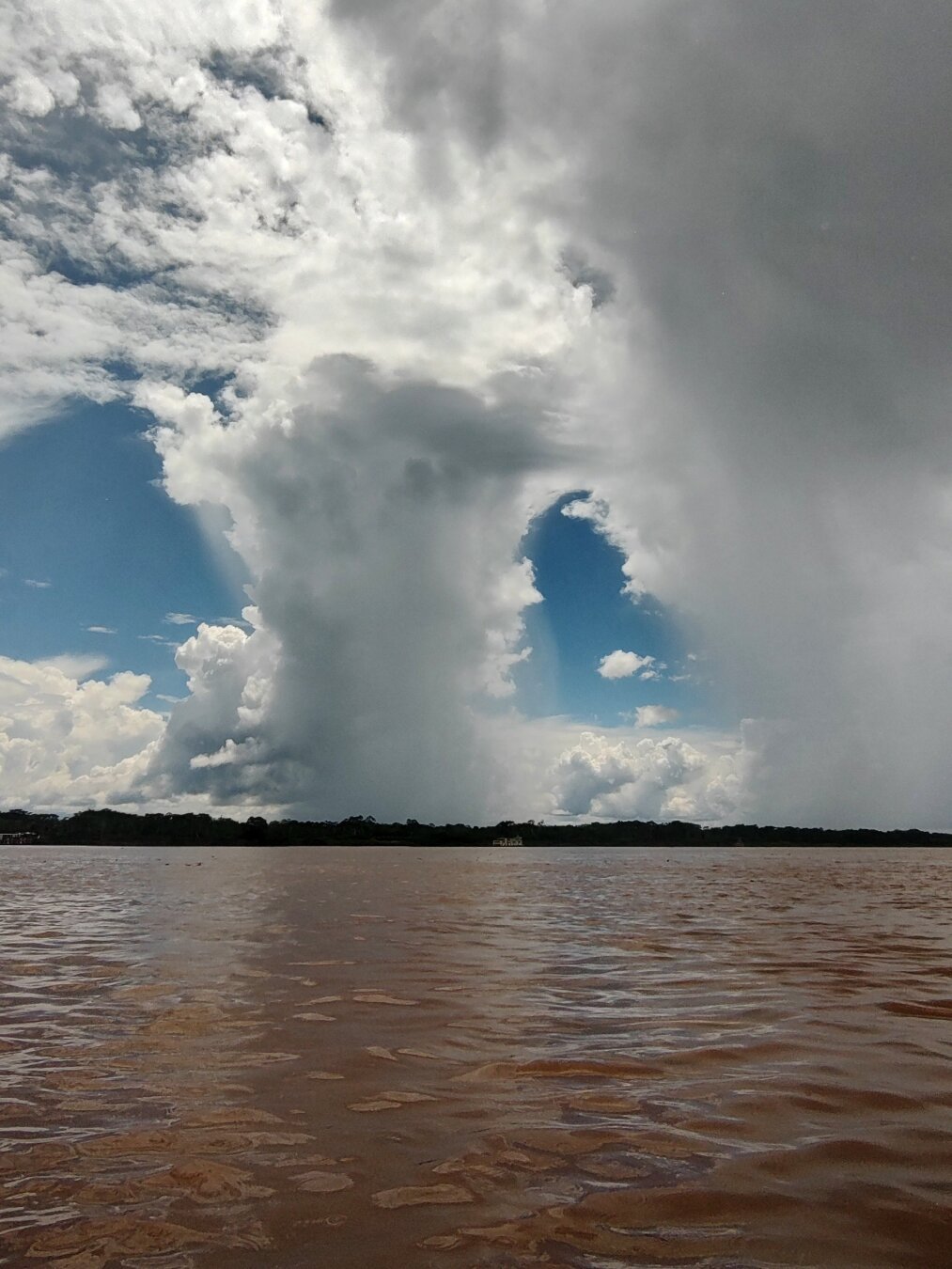 The wide, brown Amazon. In the far background, the other bank can be recognised in dark green. Absurd towers of clouds stand over the jungle and reach high into the sky. Clouds of rain fall down.