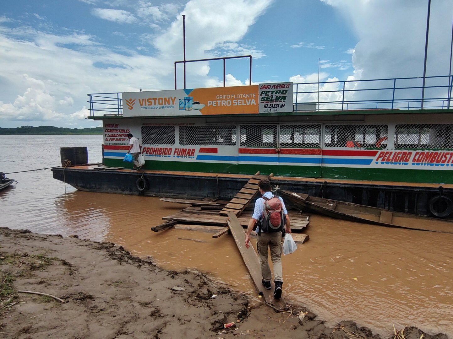 The muddy bank of the murky brown Amazon. Shaky wooden planks connect the bank with a simple ferry boat. A person with a rucksack is balancing on the wooden plank.