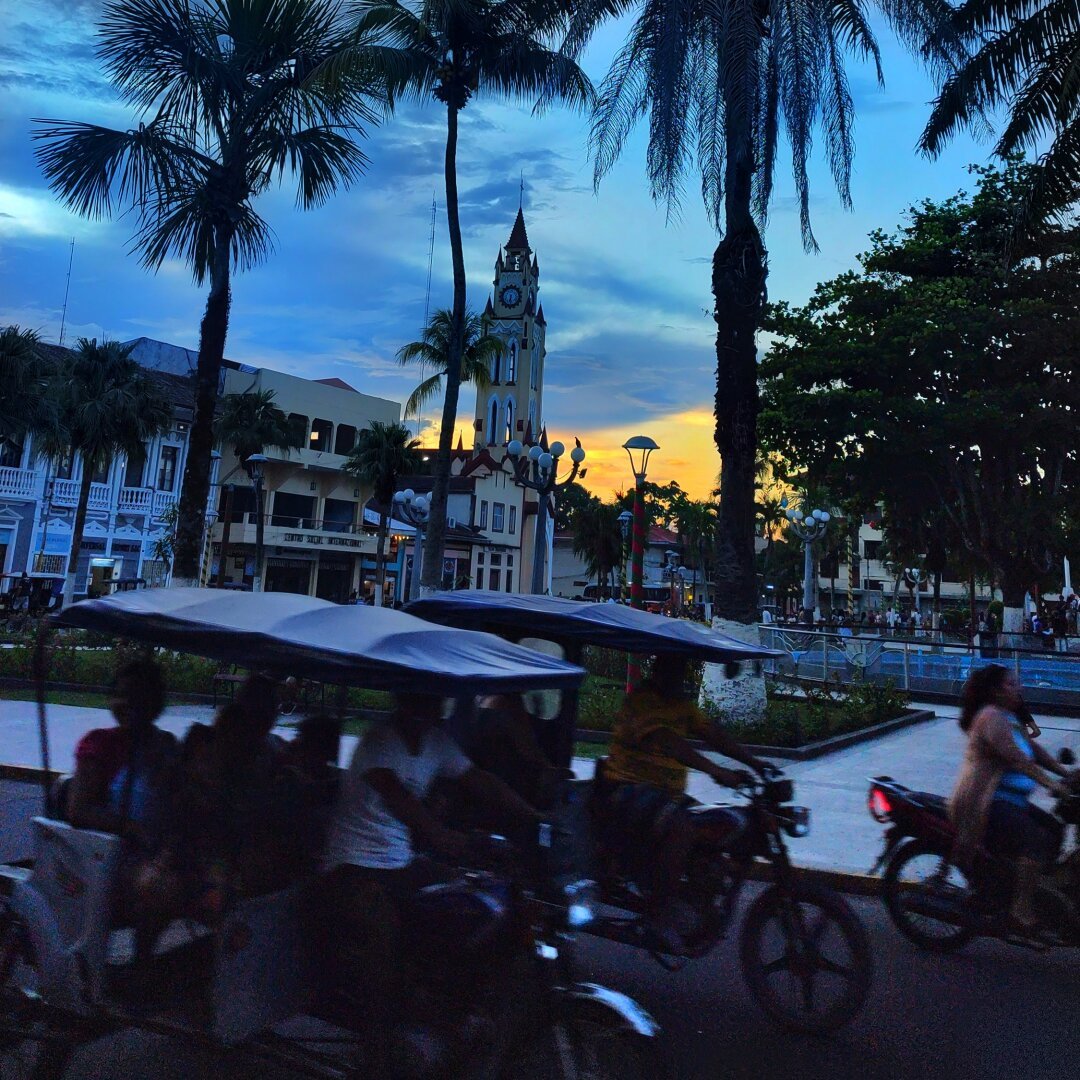 A palm-fringed square at dusk. In the background a church tower, in the foreground several motorised rickshaws. The front part is that of a moped, to which a two-seater tubular frame bench on two wheels has been welded at the back.