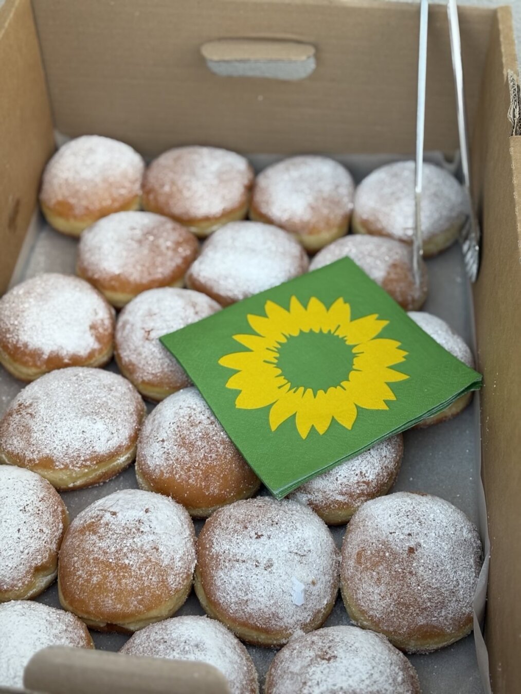 Fotografie eines Kartons, der mit Berlinern gefüllt ist. Auf einigen Berlinern liegen grüne Servietten mit der gelben Sonnenblume der Grünen
