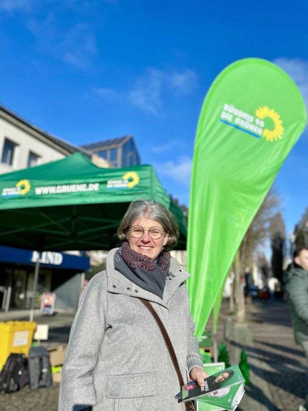 Fotografie der Bürgermeisterkandidatin  für die Kommunalwahl 2025 und Fraktionsvorsitzende der Grünen der Stadt Brühl in der Simone Holderried vor einem Wahlkampfstand der Grünen bei tollem Wetter mit strahlend blauem Himmel