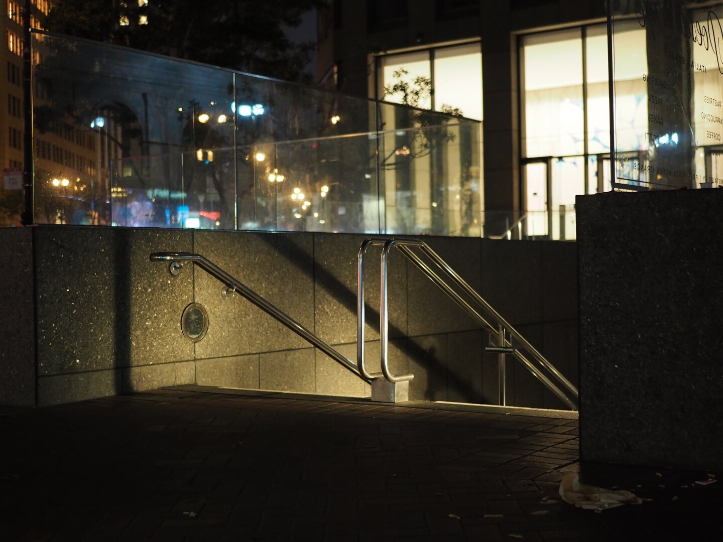 A dark street scene with stairs descending into the unknown. It is high contrast with some bright lit office windows and inky shadows. There are no people in the scene.