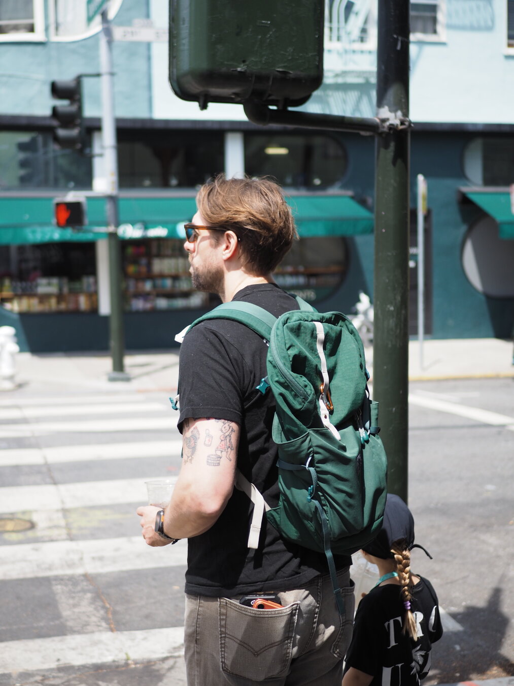 A man carrying a green backpack in front of a street light painted dark green and a store with a green awning