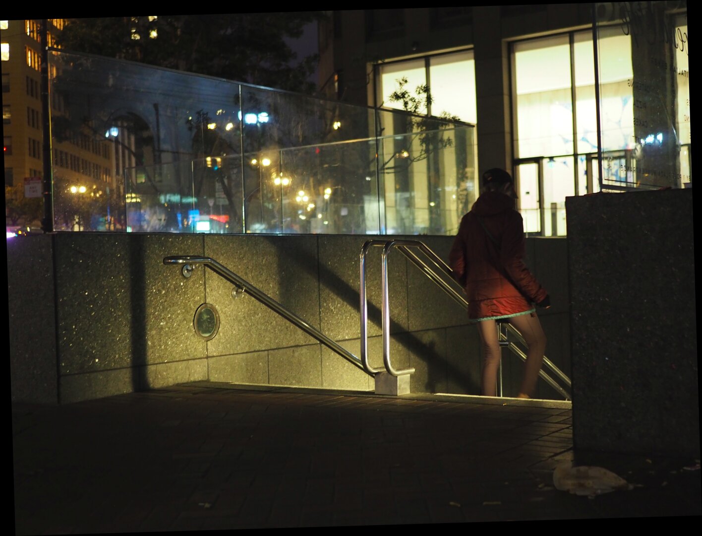 A dark street scene with stairs descending into the unknown. It is high contrast with some bright lit office windows and inky shadows. A flitting shadowy figure descents the stairs.