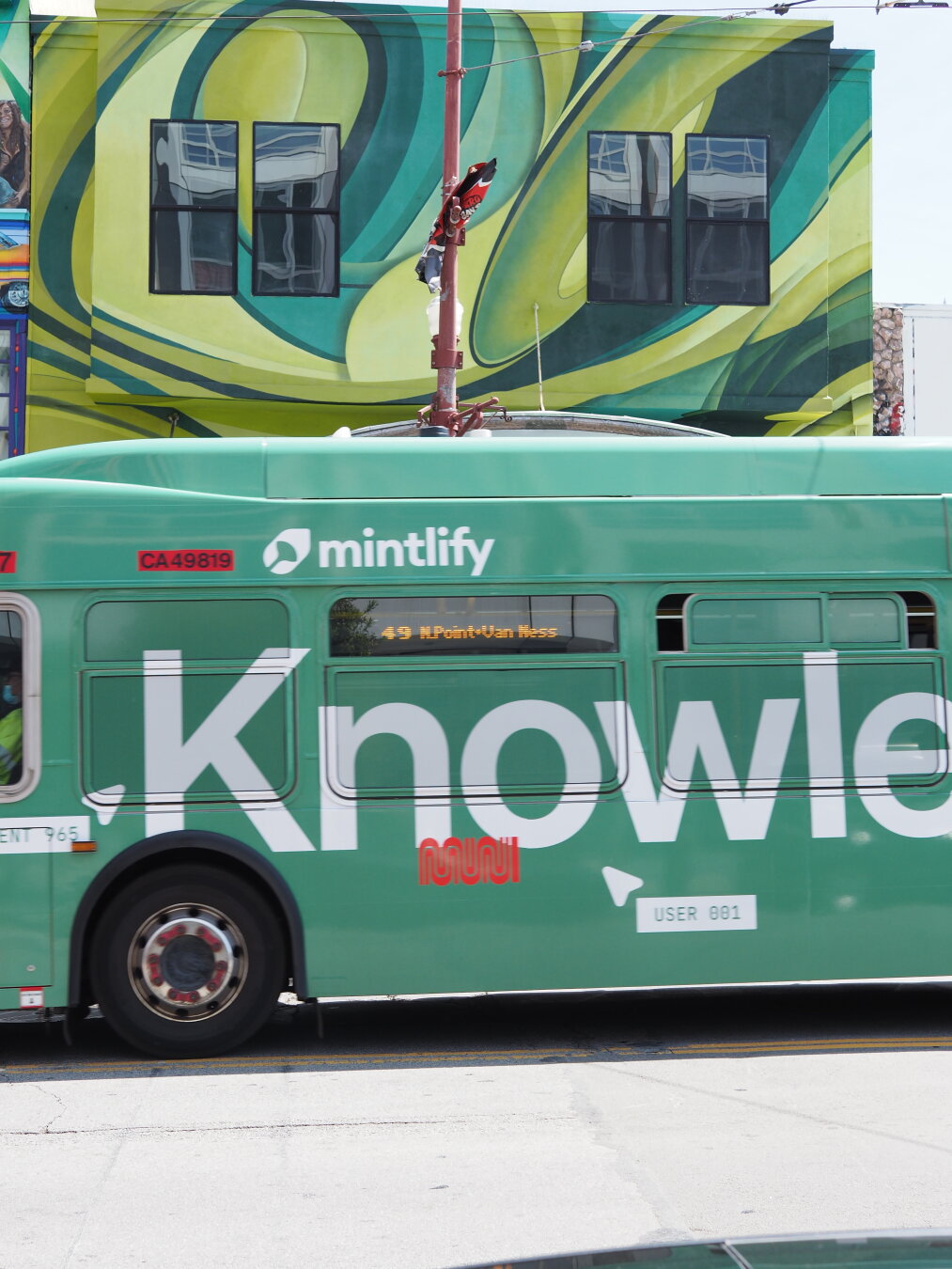 A green SF Muni bus with a green "Knowledge" ad wrapper passing a building painted with bright green leaves