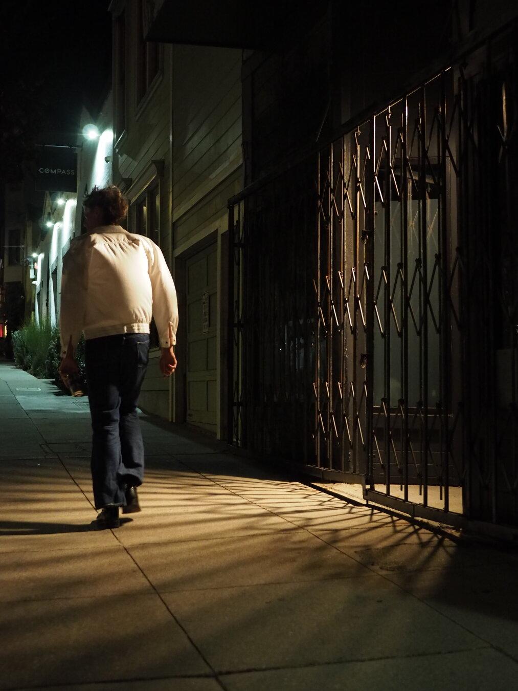 A man in a white jacket walks through a pool of light streaming through a security gate. The wind is making his jacket billow out, and the light emphasizes its curving shape.