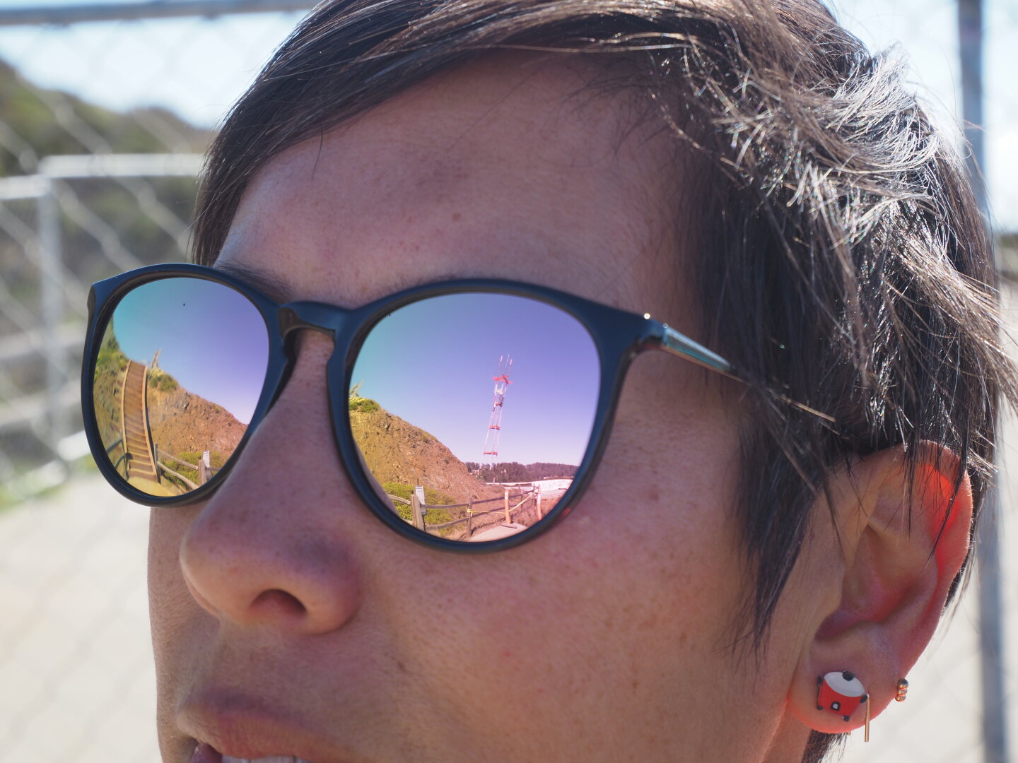A close-up photo of a woman's face. She's wearing reflective sunglasses. In the left lens a staircase is reflected, and in the right lens a red and white tower is reflected.