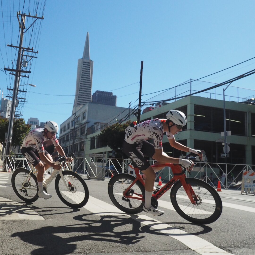 Two bicycle racers lean over as they chase each other around a corner on a city street. In the background, the Transamerica Pyramid looms