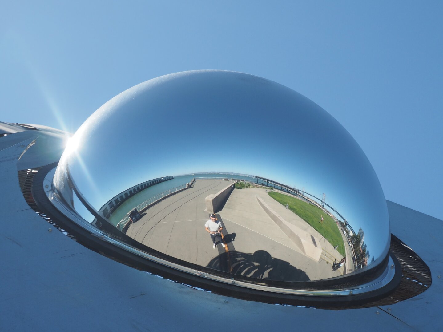 A self portrait in the curved and reflective eye of a fish sculpture on the Embarcadero in San Francisco. The photo shows the photographer in the foreground with a park and Bay Bridge in the background. The further from the mirror, the greater the curving distortion