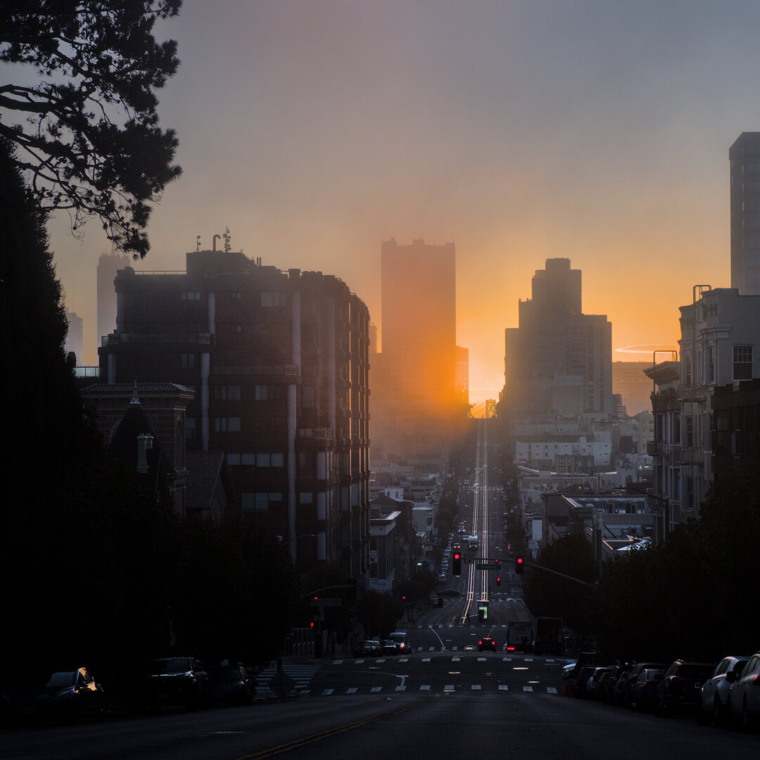 A photo looking down California Street from Gough Street, with the sunrise in the gap between the tall buildings in the distance. The top of the Bay Bridge can be seen. The sunlight is reflecting on the cablecar tracks, whichs shine brightly against the dark street.
