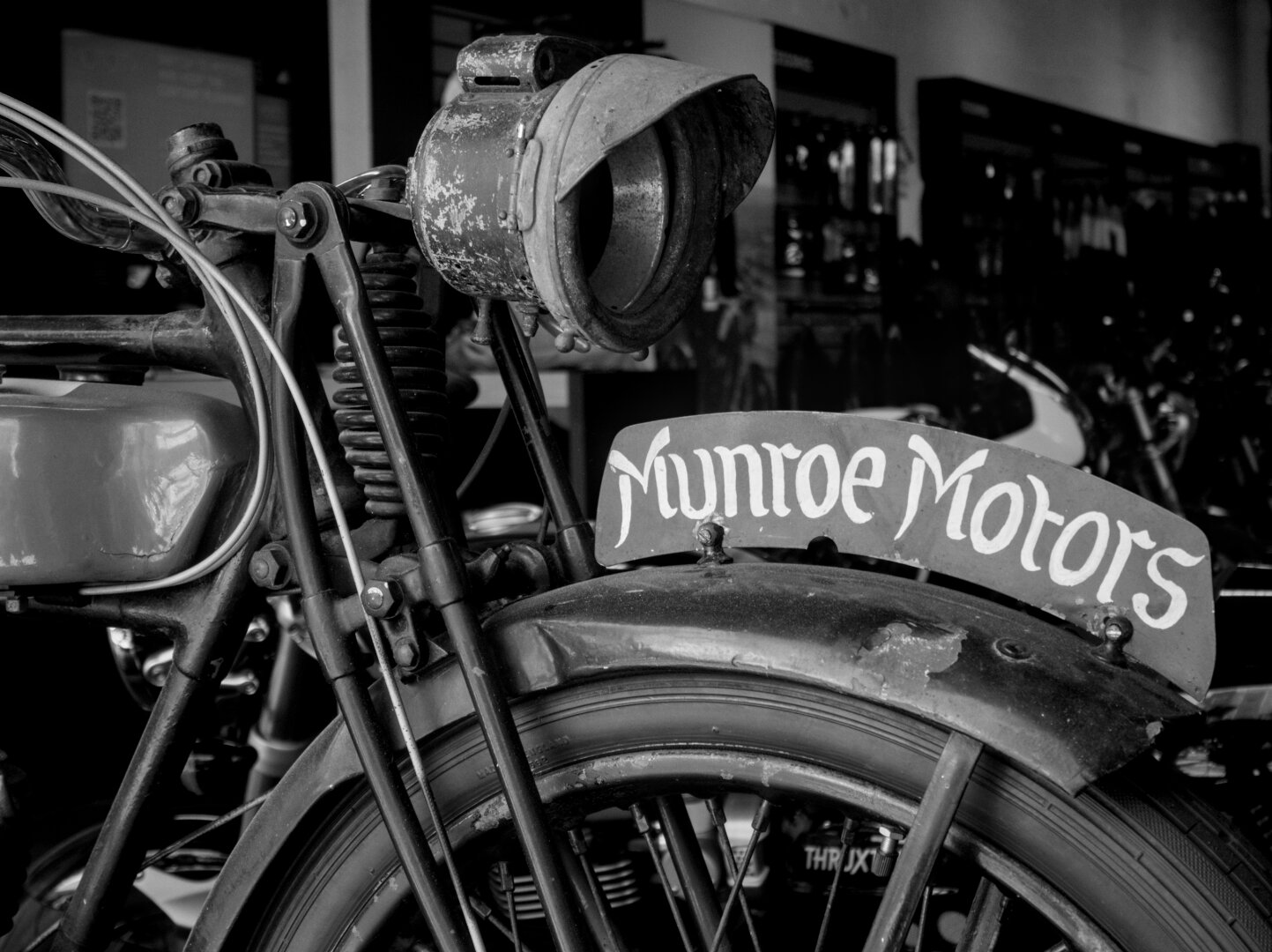 A black and white shot of a vintage Monroe Motors motorbike. The shot includes the front of the gas tank, a springer-style fork, a weathered lamp, and a metal fender with a sign attached reading "Monroe Motors"