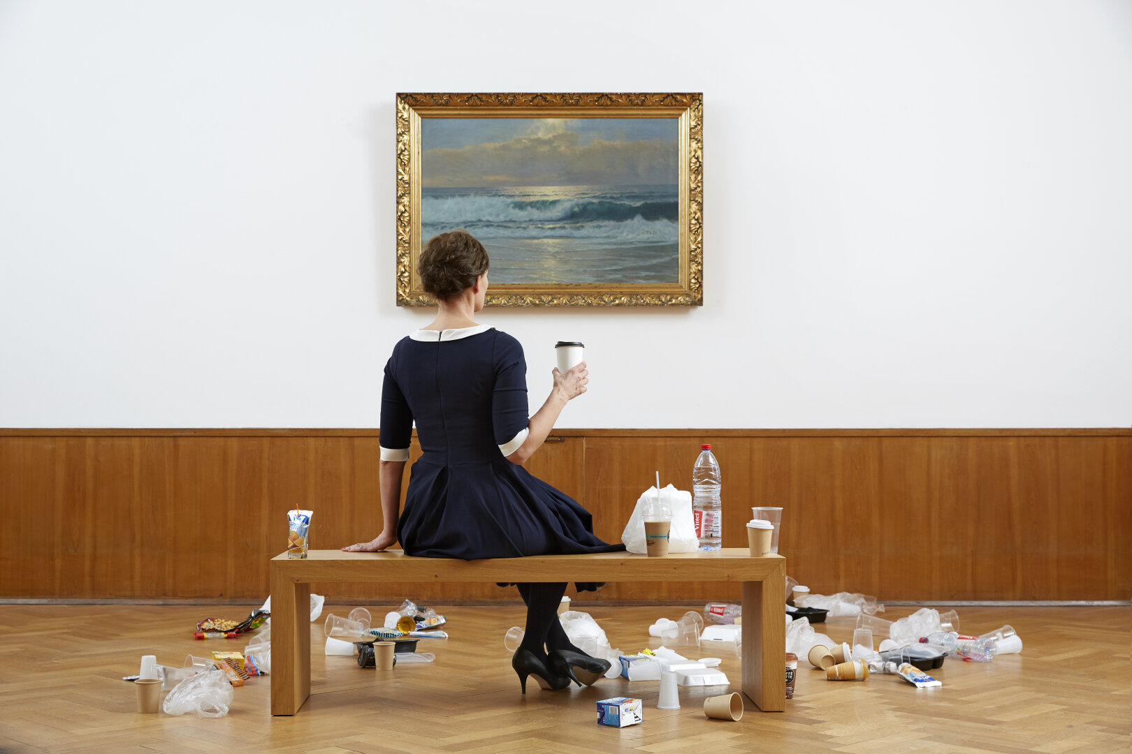 The artist Swaantje Güntzel sits on a museum bench in front of a seascape depicting the surf of the North Sea.  She has her back to the viewer and is wearing a blue and white navy dress. She is holding a coffee-to-go cup in her hand. Around her, on the bench and on the floor of the room, lie various disposable plastic packaging items such as bags, takeaway boxes, etc.