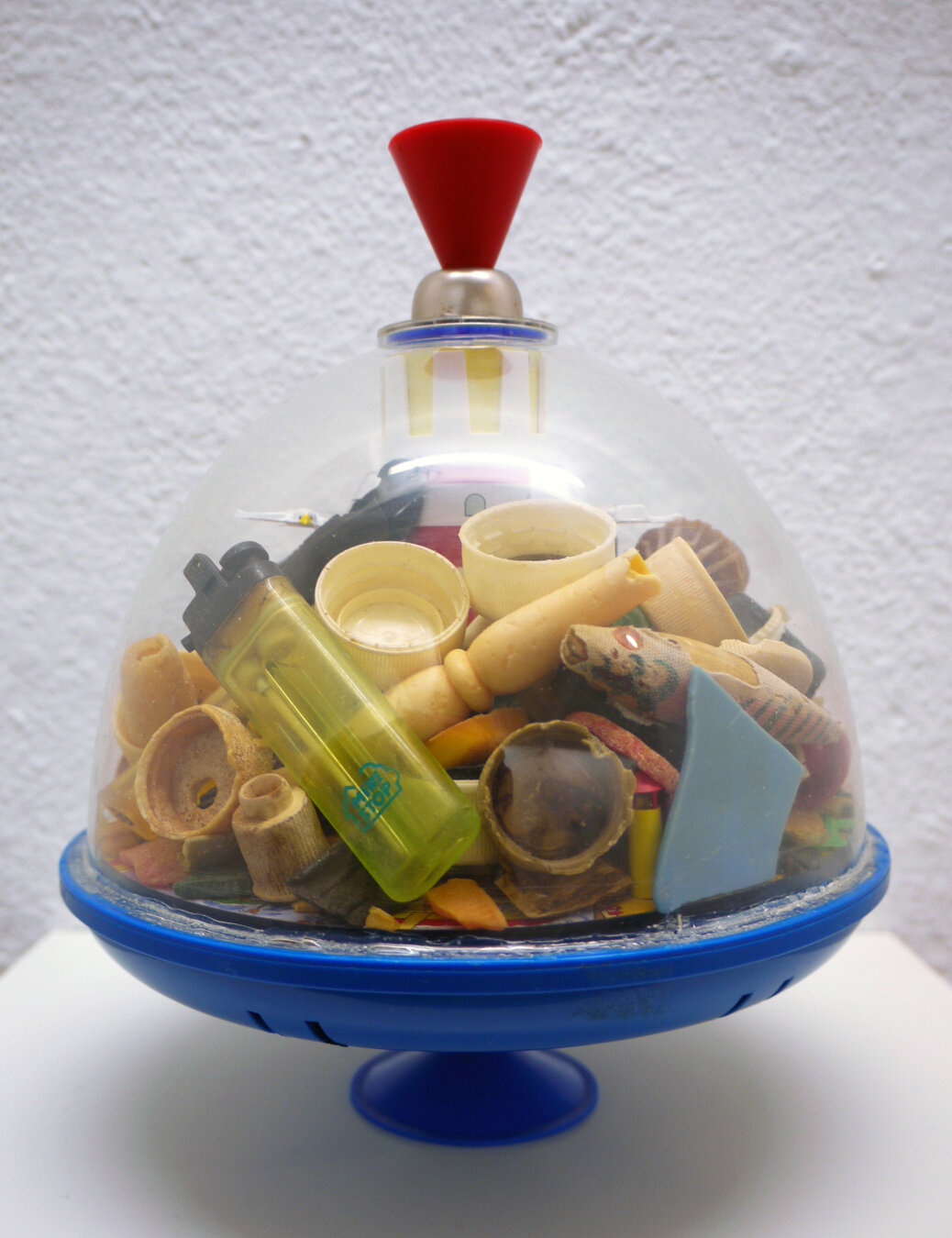 The image shows a toy spinning top for children with a blue base and red handle. Inside the spinning top are various plastic objects, such as lighters, bottle caps and toys. The objects were found in the stomach of an albatross chick on Kure Atoll.