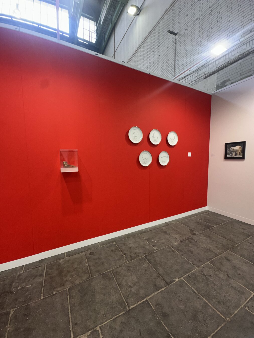 Red wall displaying several art works of the artist Swaantje Güntzel at Postions Art Fair in Berlin. We see 5 white porcelain plates, a transparent small acrylic box with a brinze hand and on te right side a framed image of a  polar bear.