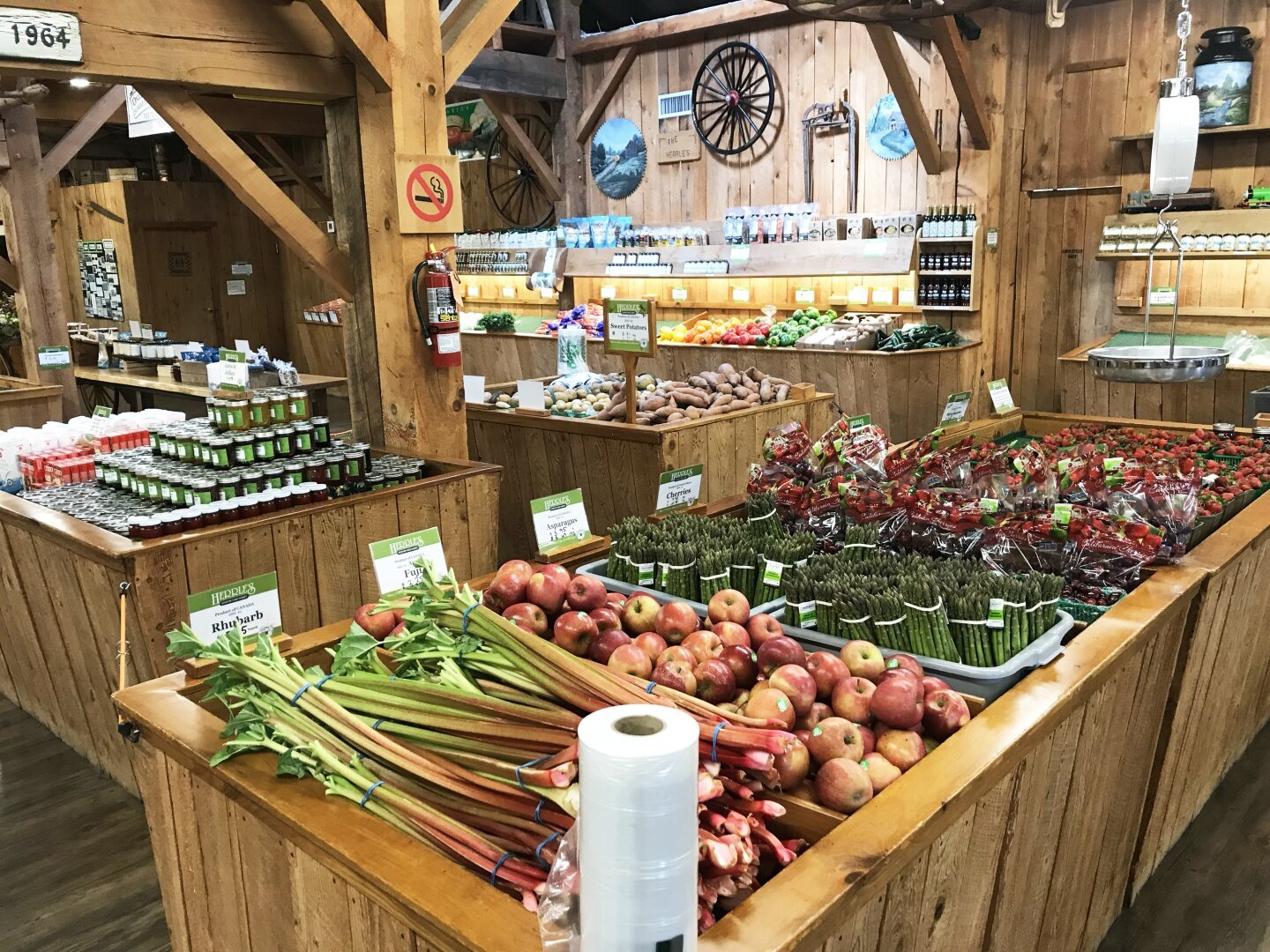 The interior of Herrles. Bins of rhubarb, apples, asparagus, berries, strawberries are in the foreground. In the background are courgettes, mushrooms, red, green, and gold capsicum peppers, carrots, sweet potatoes. There's also another row of bins with canning and canning supplies. The walls are barnboard with various jugs and wheels and things.