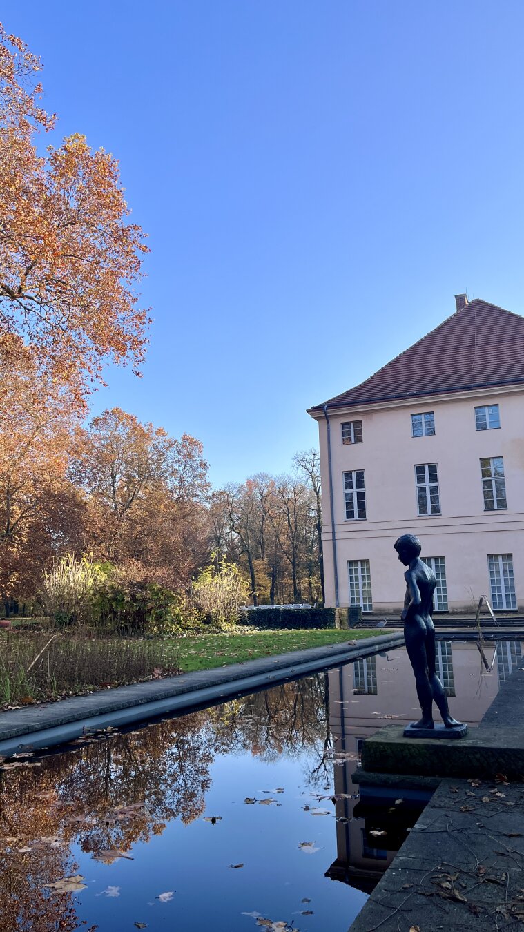 Bronze statue of a boy by a reflecting pool in front of Schloss Niederschönhausen on a clear autumn day.