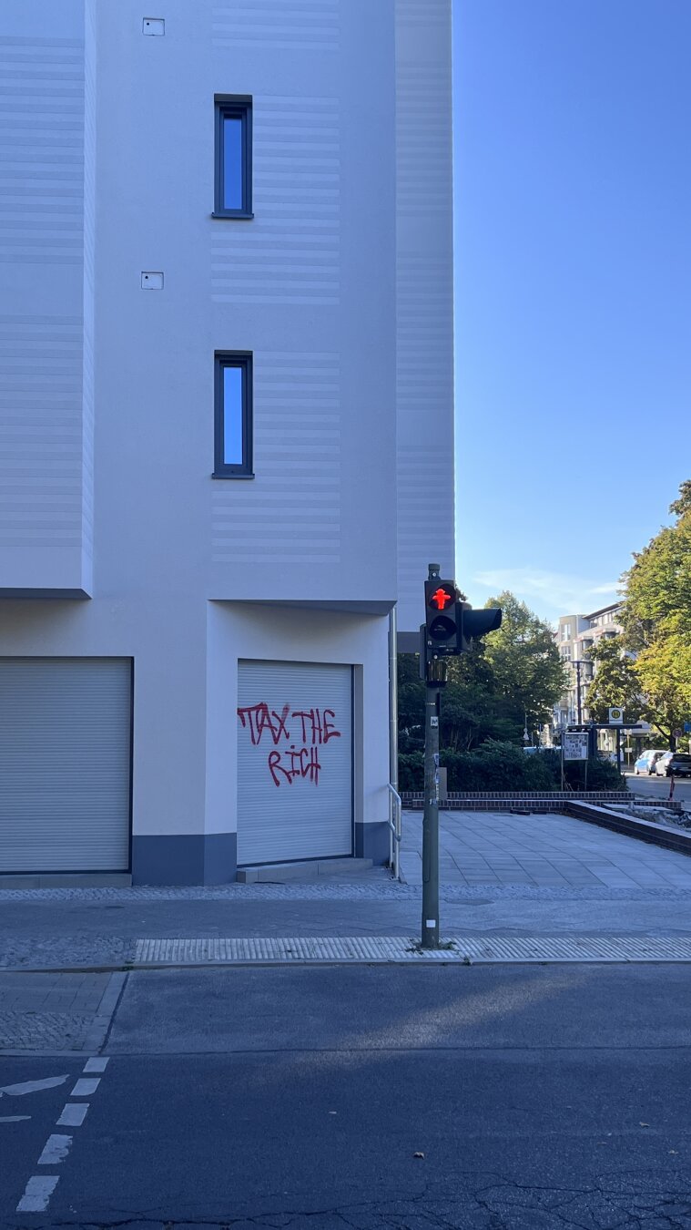 White new building with red graffiti reading “TAX THE RICH” and a red pedestrian stop light.