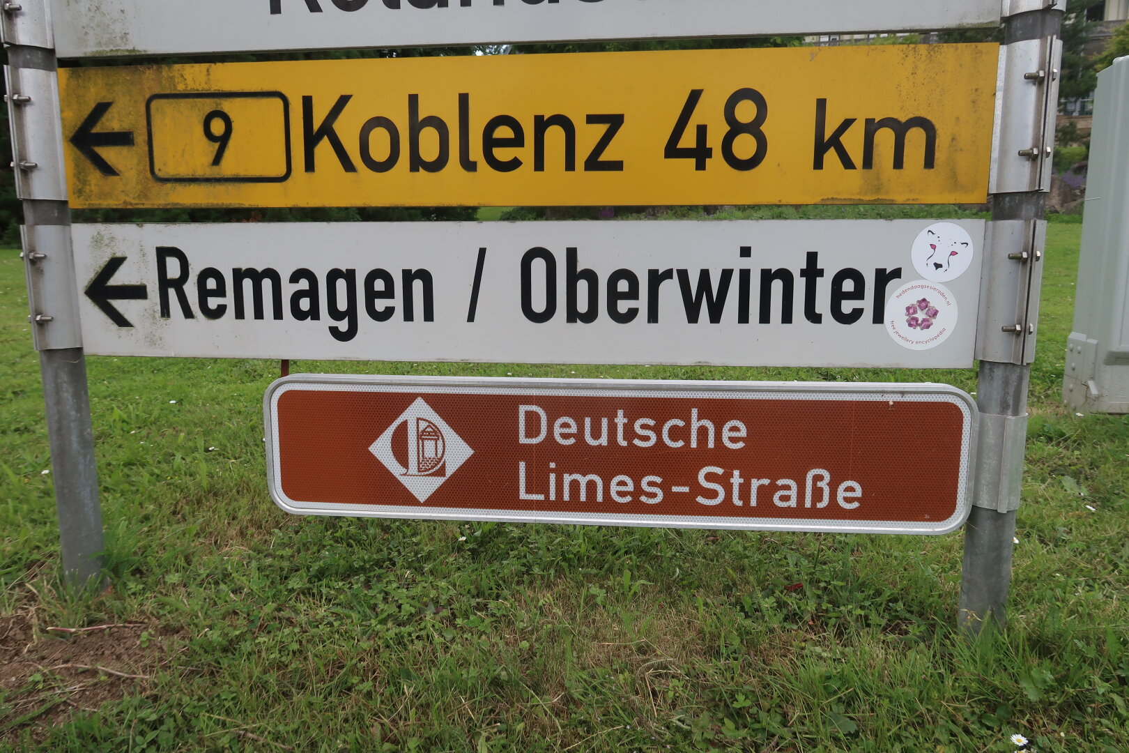 Ronde sticker van hedendaagsesieraden.nl onder ronde sticker met kop van katachtige op plaatsnaamborden in Duitsland.